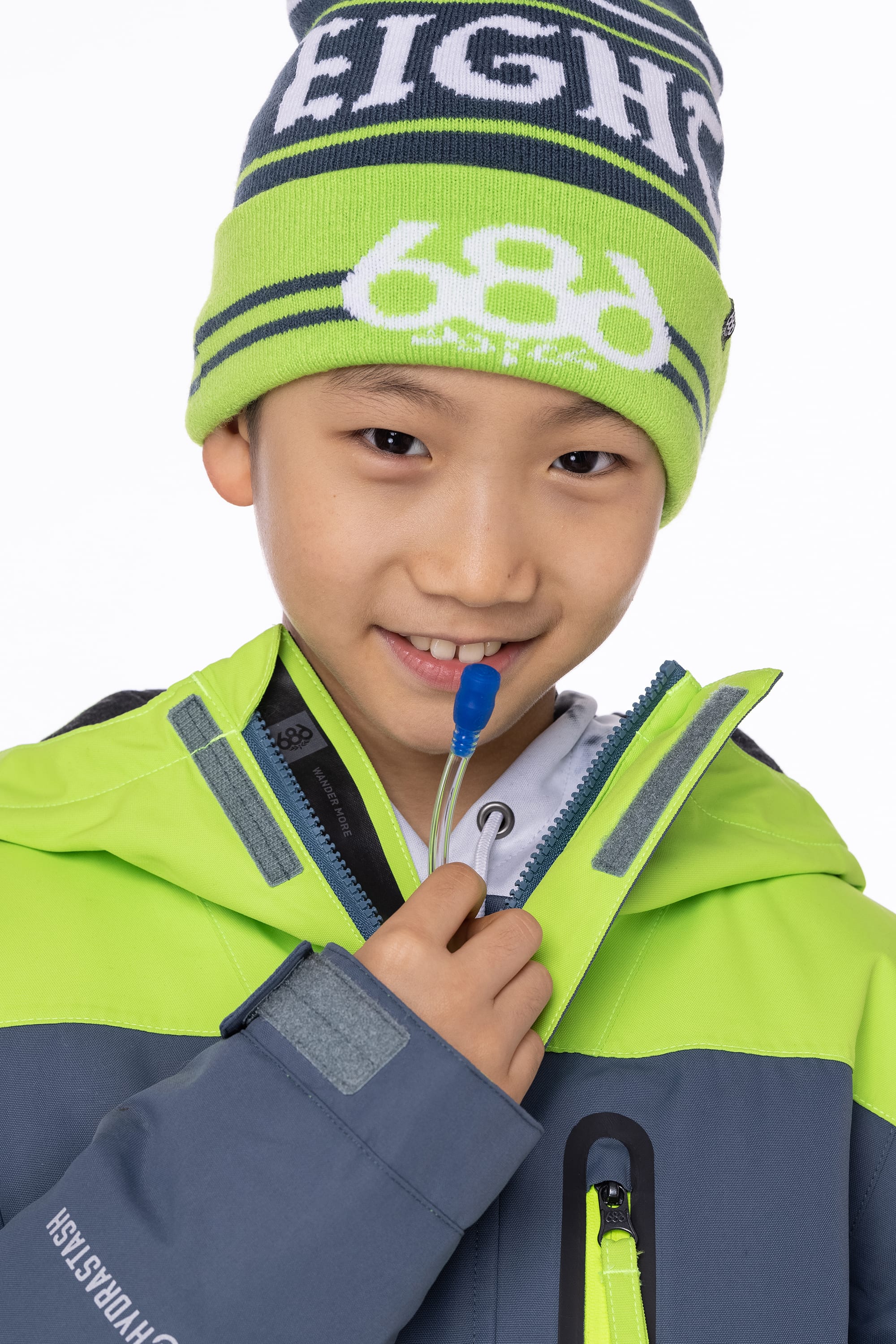 A smiling child in the 686 Boys Hydrastash Insulated Jacket and beanie holds a blue zipper pull to their mouth, looking directly at the camera against a white background.