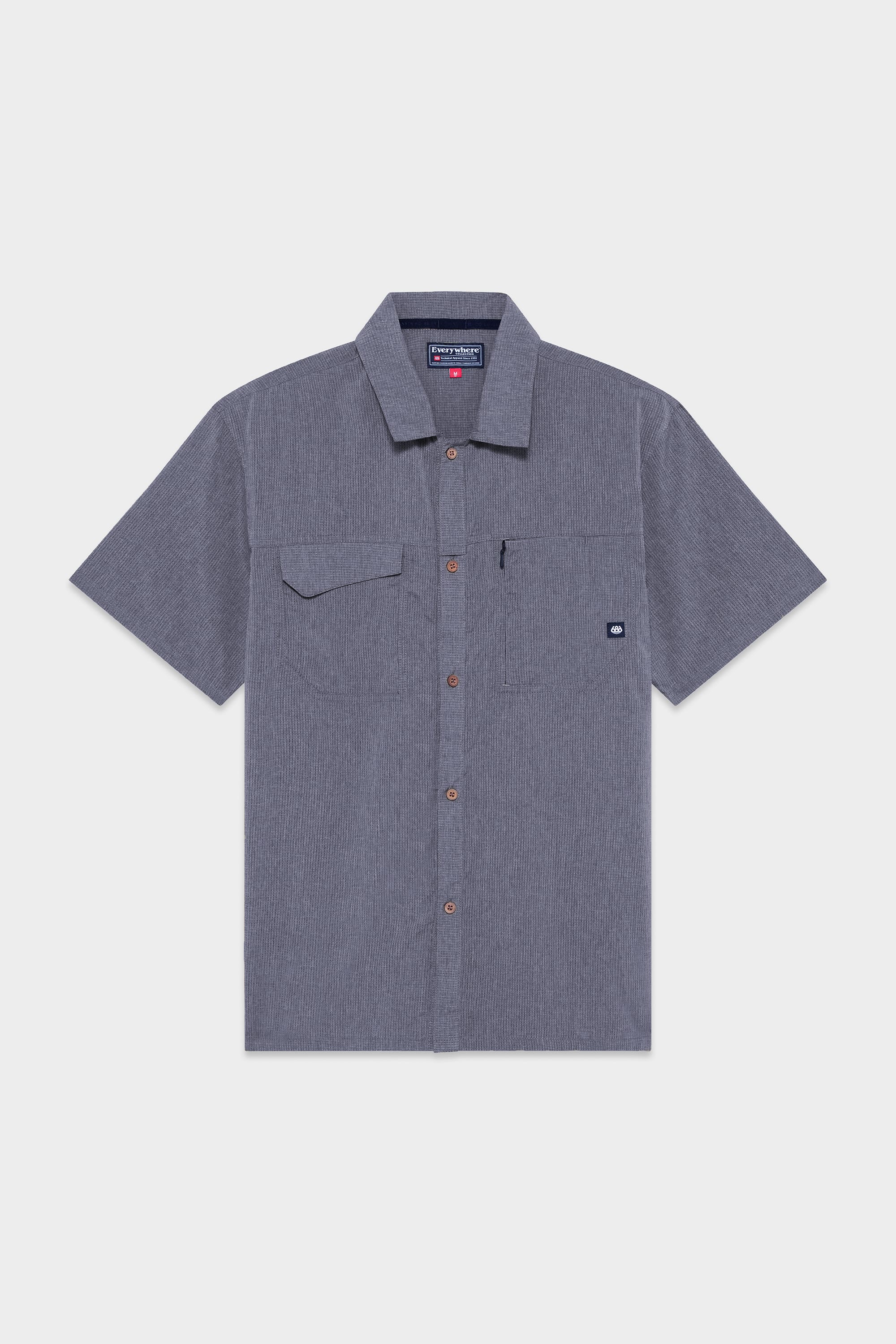 The 686 Mens Seeker Perforated Button Up features short sleeves, a collar, brown buttons, a left chest pocket with a brand logo tag, and a subtle textured pattern in grey.