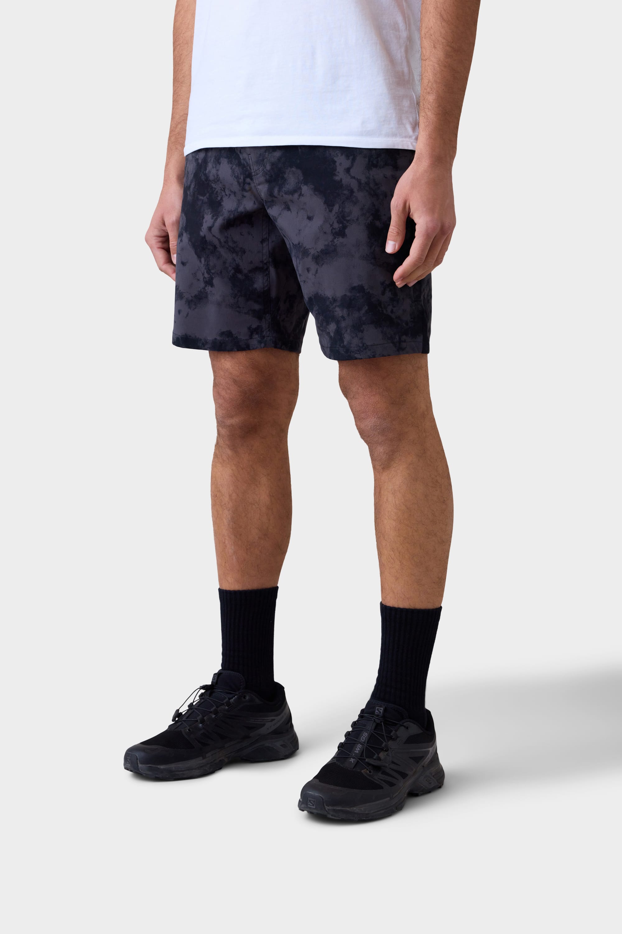 A person stands against a plain light background, wearing a white T-shirt, black 686 Mens Everywhere Hybrid Short, black crew socks, and black sneakers. The stylish, stain-resistant hybrid shorts from 686 are breathable. Their face is not visible.