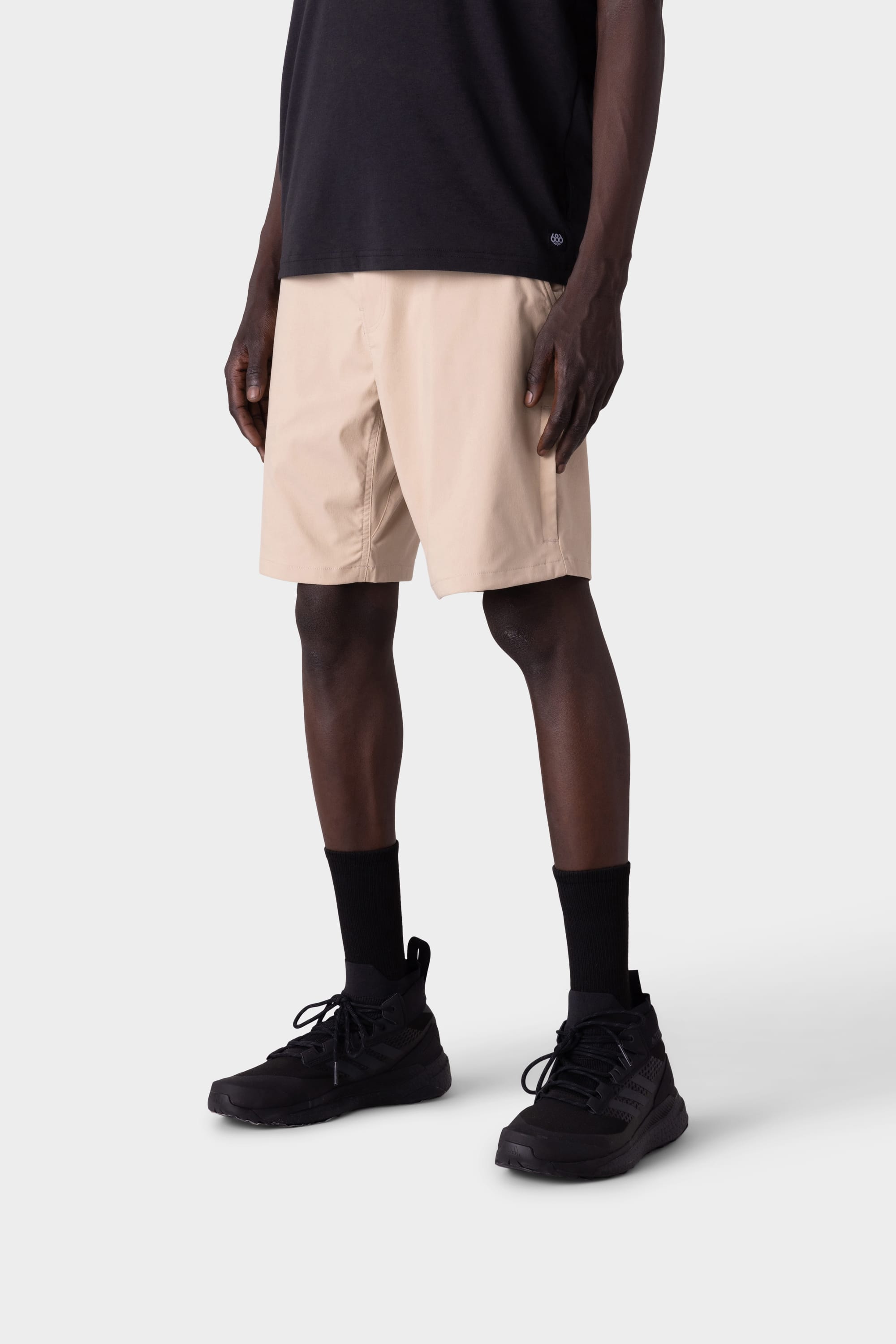 A person wearing a black t-shirt, 686 Mens Everywhere Hybrid Short by 686, black crew socks, and black sneakers stands against a light gray background. The breathable, stain-resistant shorts are shown from shoulders to feet.