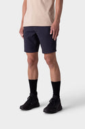A person wears a beige breathable t-shirt, dark navy 686 Mens Everywhere Hybrid Short, black crew socks, and black sneakers, standing against a plain light gray background. The photo is cropped from the shoulders down.
