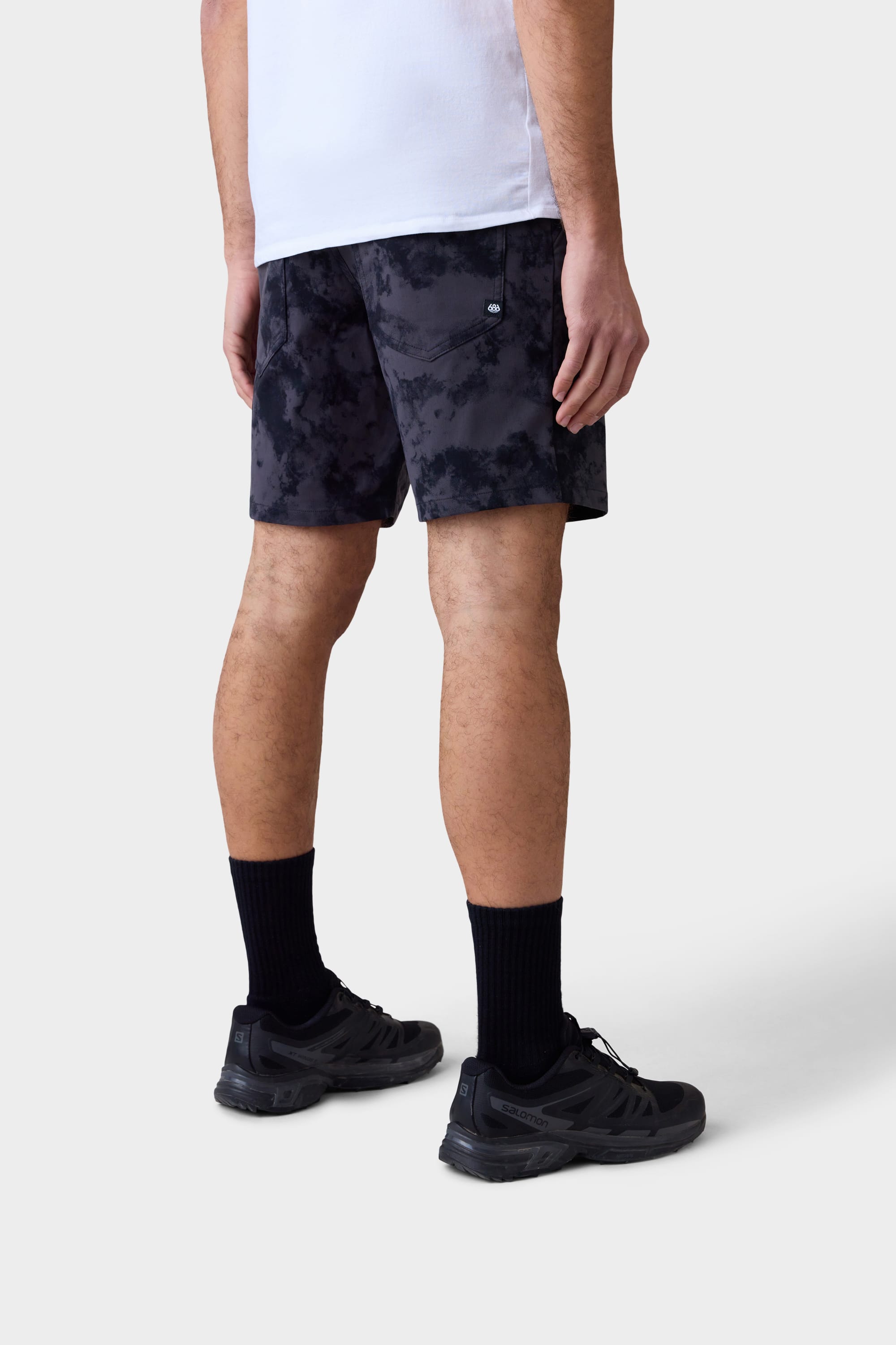 A person stands against a plain background, shown from the waist down and facing slightly away, wearing a white shirt, black 686 Mens Everywhere Hybrid Short, black crew socks, and black athletic sneakers.