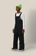 A young person wears the 686 Boys Frontier Insulated Bib snow pants with light insulation, a white long-sleeve shirt, black boots, and a neon green knit beanie, standing against a plain light background and smiling.