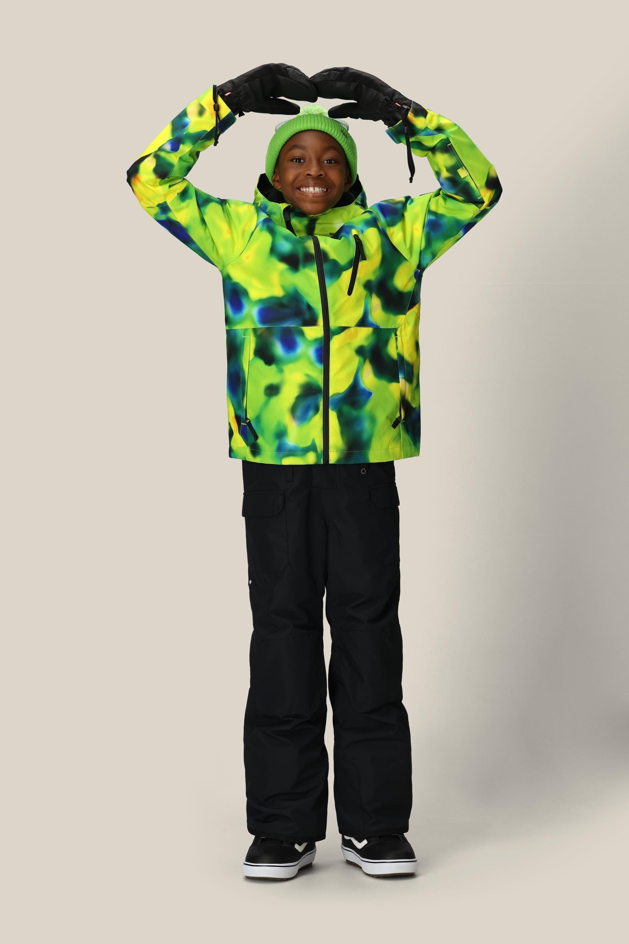A smiling child in the 686 Boys Frontier Insulated Bib, paired with a colorful jacket, black gloves, and a green beanie, stands with arms raised and hands forming a heart shape overhead.