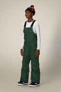 A boy wearing a white long-sleeve shirt, 686 Boys Frontier Insulated Bib in dark green, black and white sneakers, and a black beanie with red trim smiles against a plain light background.