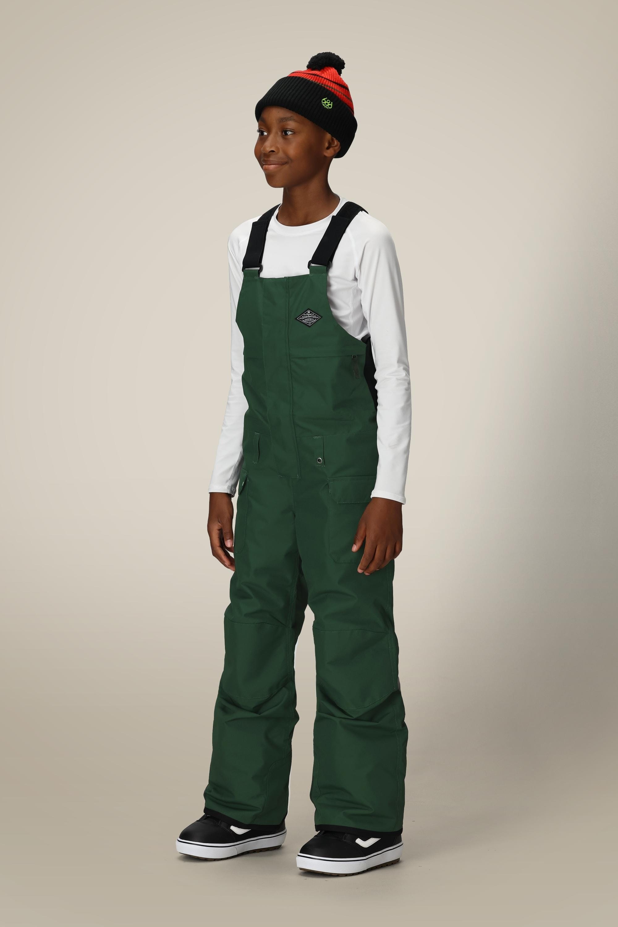 A boy wearing a white long-sleeve shirt, 686 Boys Frontier Insulated Bib in dark green, black and white sneakers, and a black beanie with red trim smiles against a plain light background.