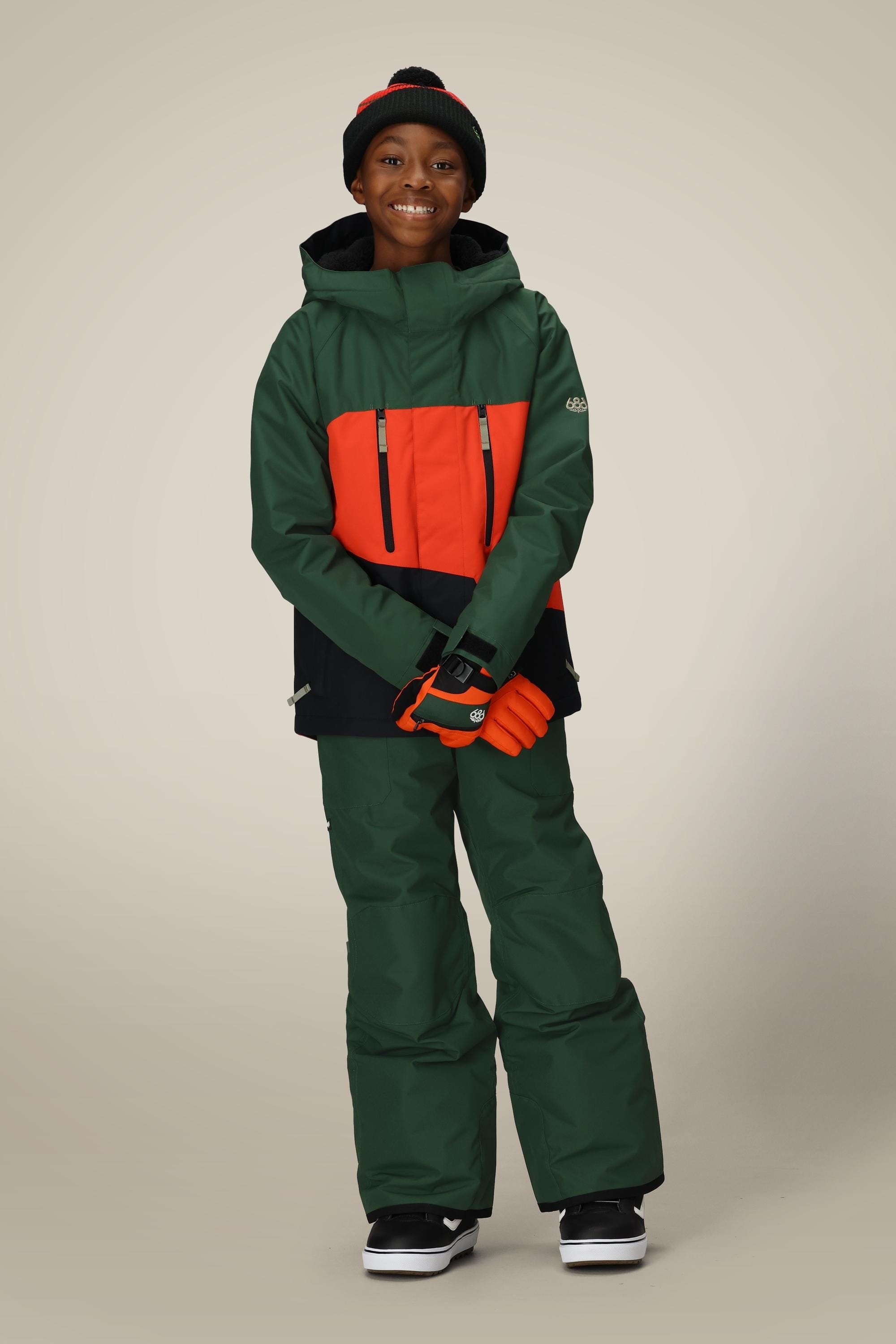 A smiling child stands against a plain background wearing the 686 Boys Frontier Insulated Bib, a green and orange winter jacket, orange gloves, black and red beanie, and black sneakers.