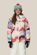 A person wears the 686 Girls Athena Insulated Jacket by 686 with white snow pants, black gloves, and a light green knit beanie with a pink pom-pom, smiling against a plain beige background.