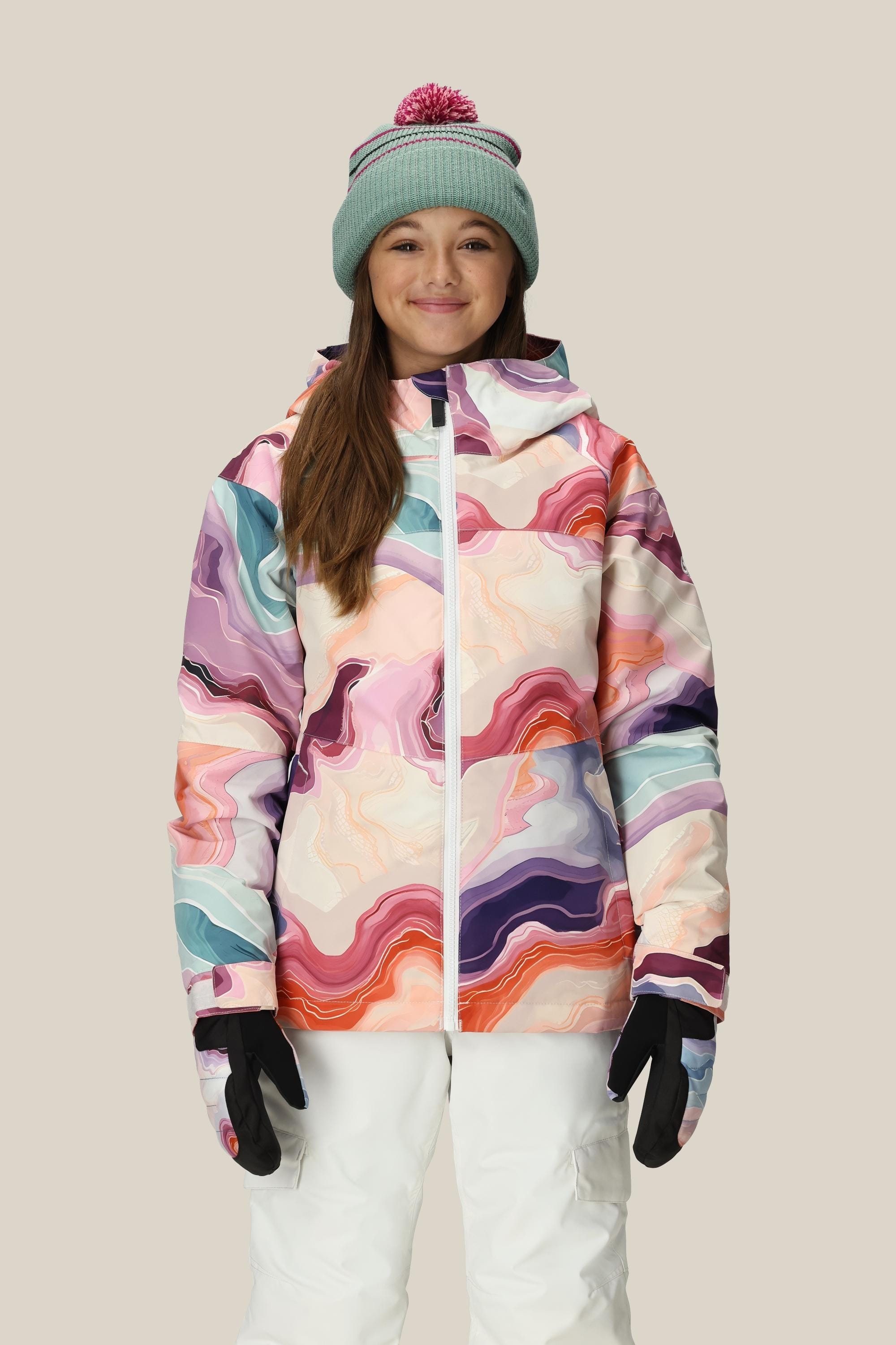 A person wears the 686 Girls Athena Insulated Jacket by 686 with white snow pants, black gloves, and a light green knit beanie with a pink pom-pom, smiling against a plain beige background.