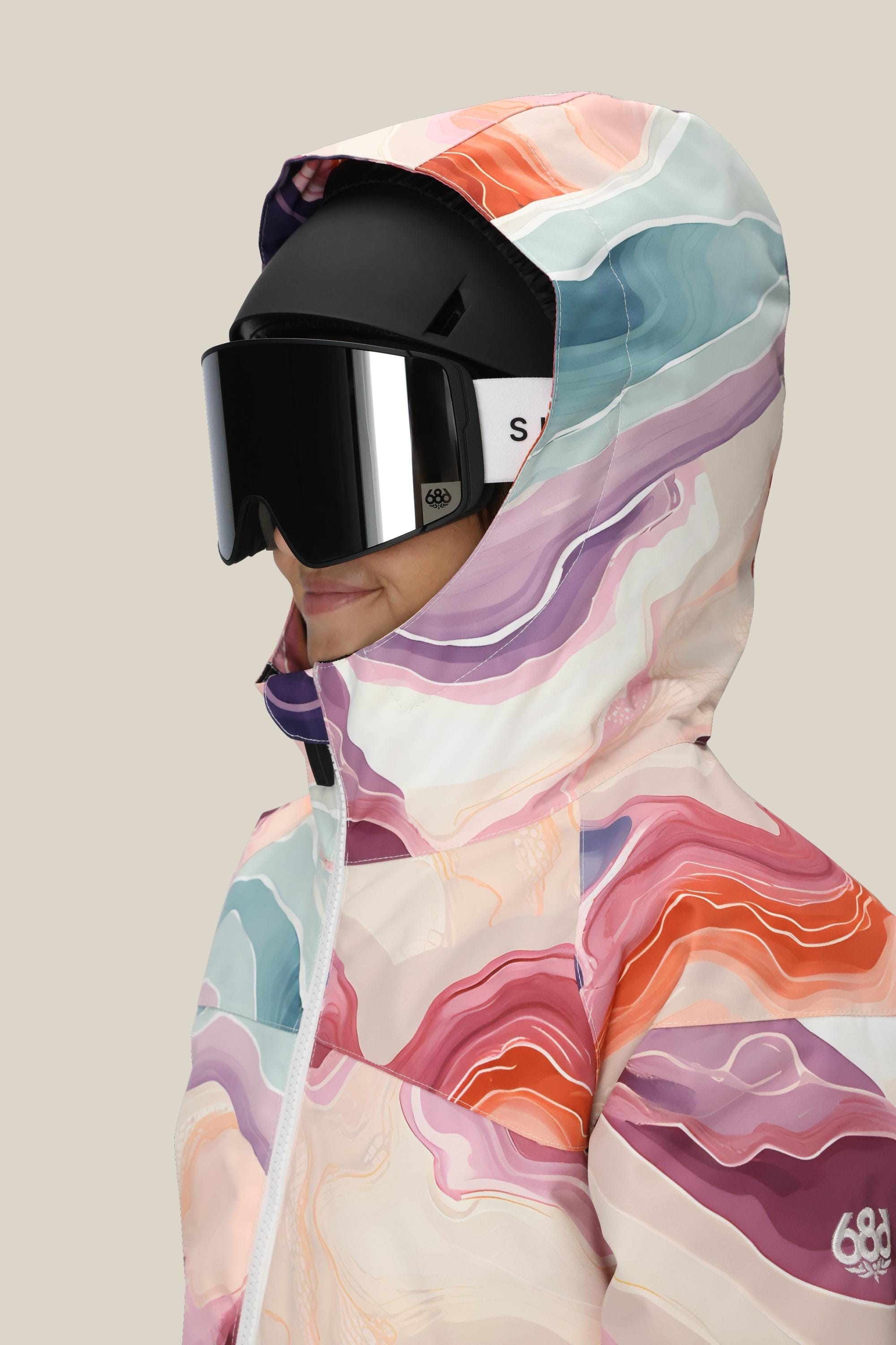 A girl wears the 686 Girls Athena Insulated Jacket—multicolored with a marble pattern—paired with a black ski helmet and large black goggles, shown in profile against a plain beige background.