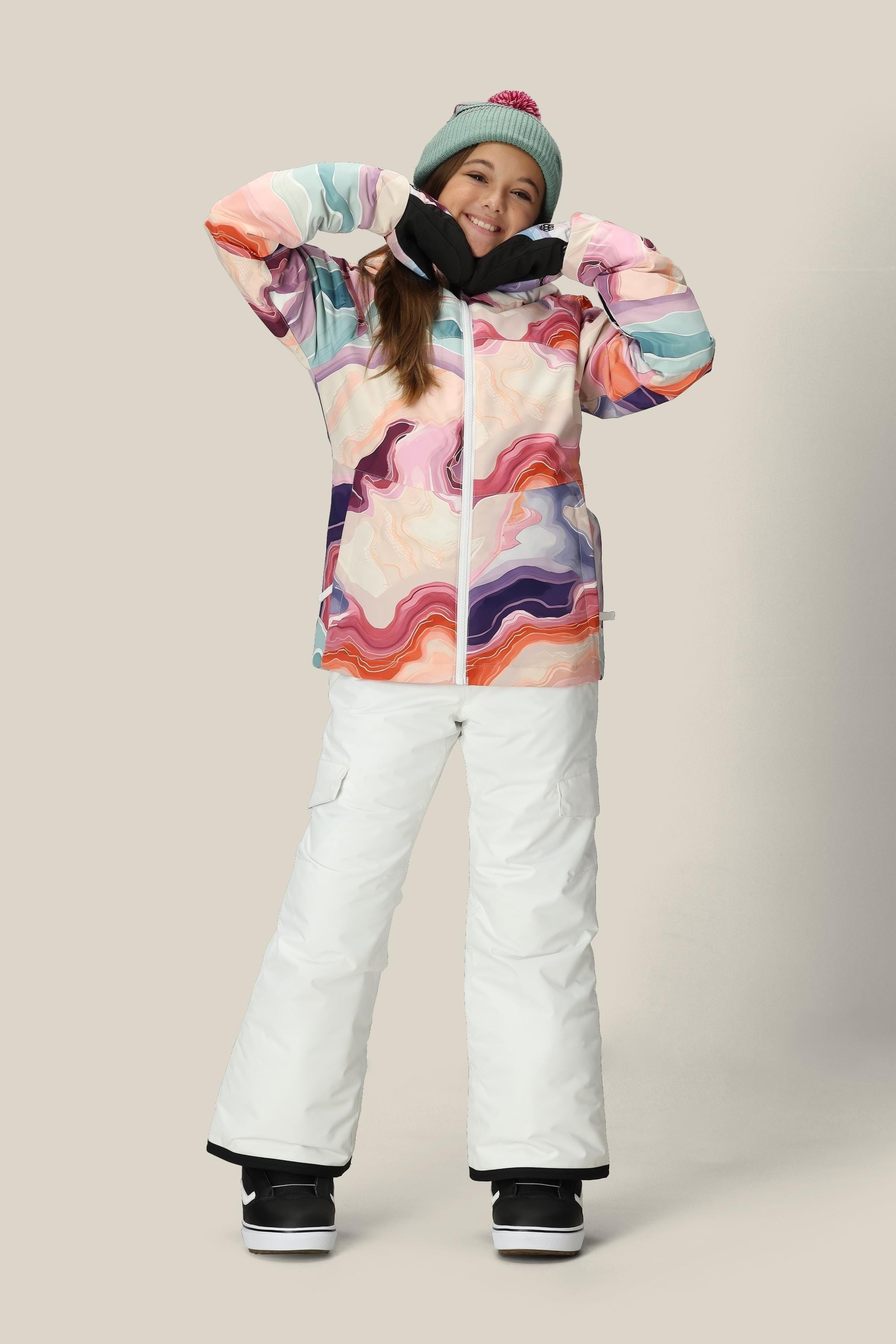 A smiling person wears the 686 Girls Athena Insulated Jacket by 686, featuring a colorful abstract pattern, along with white snow pants, black gloves, snow boots, and a green beanie with a pink pom-pom against a plain light background.