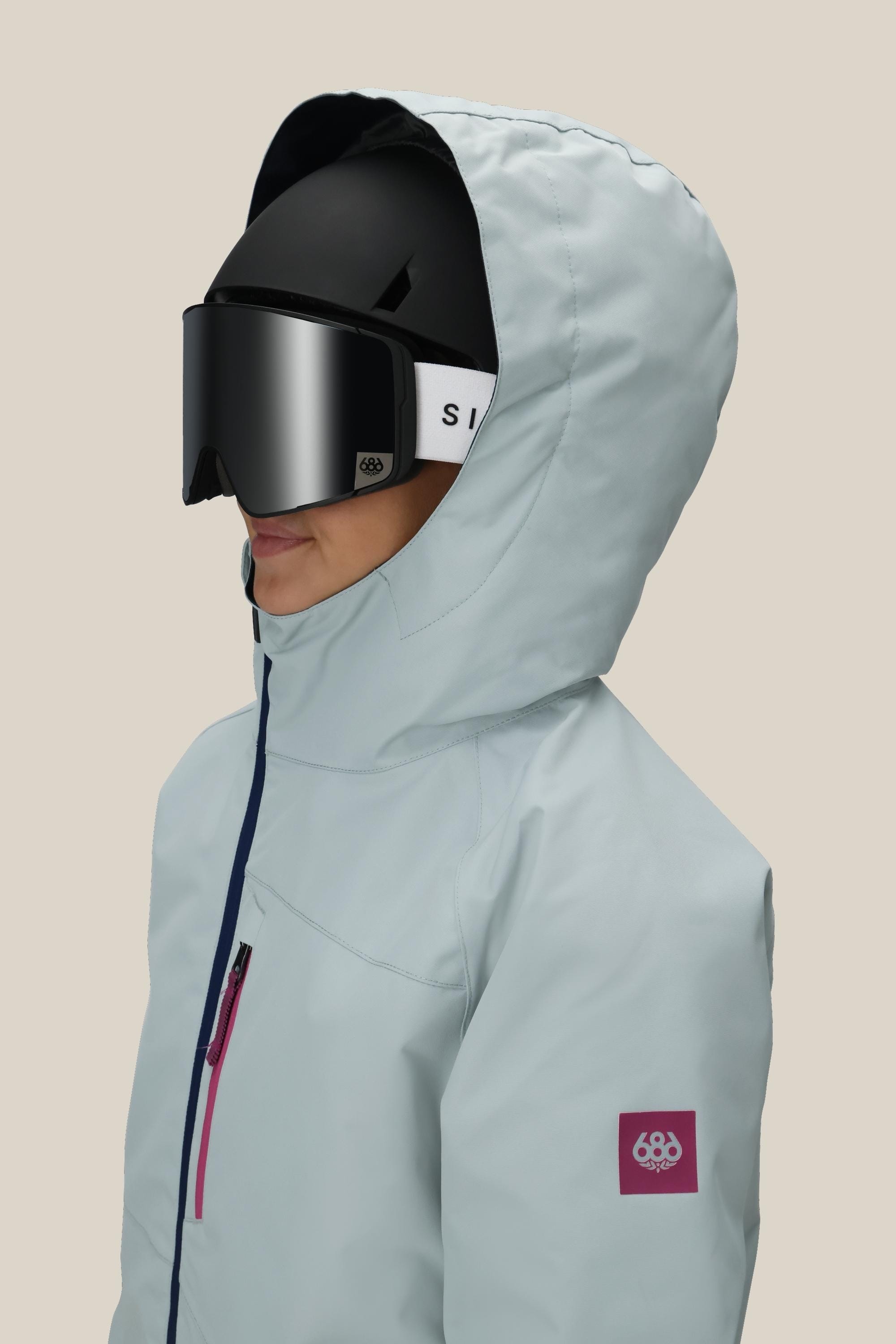 A girl in profile wears the 686 Girls Hydra Insulated Jacket in light blue, paired with a black ski helmet and black goggles with white straps, set against a plain beige background.