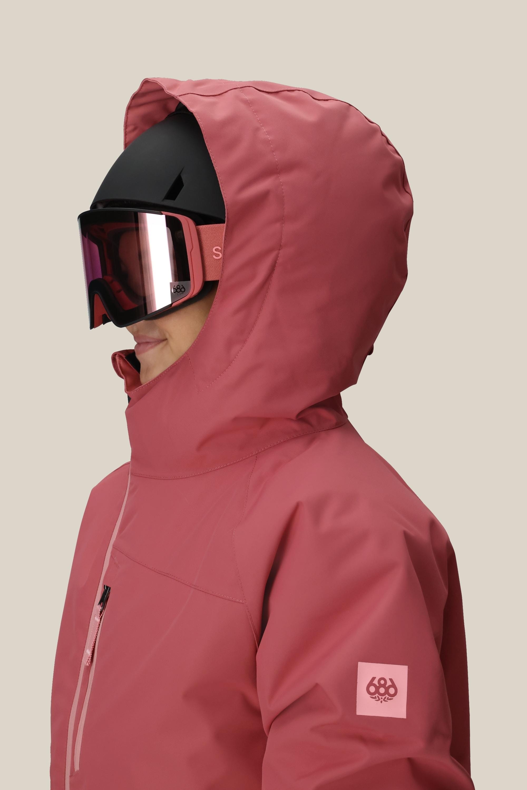 A person in the 686 Girls Hydra Insulated Jacket, featuring body-mapped lining for comfort and performance, is shown in profile. The best-selling pink jacket has a small arm logo and is paired with a black ski helmet and reflective goggles.
