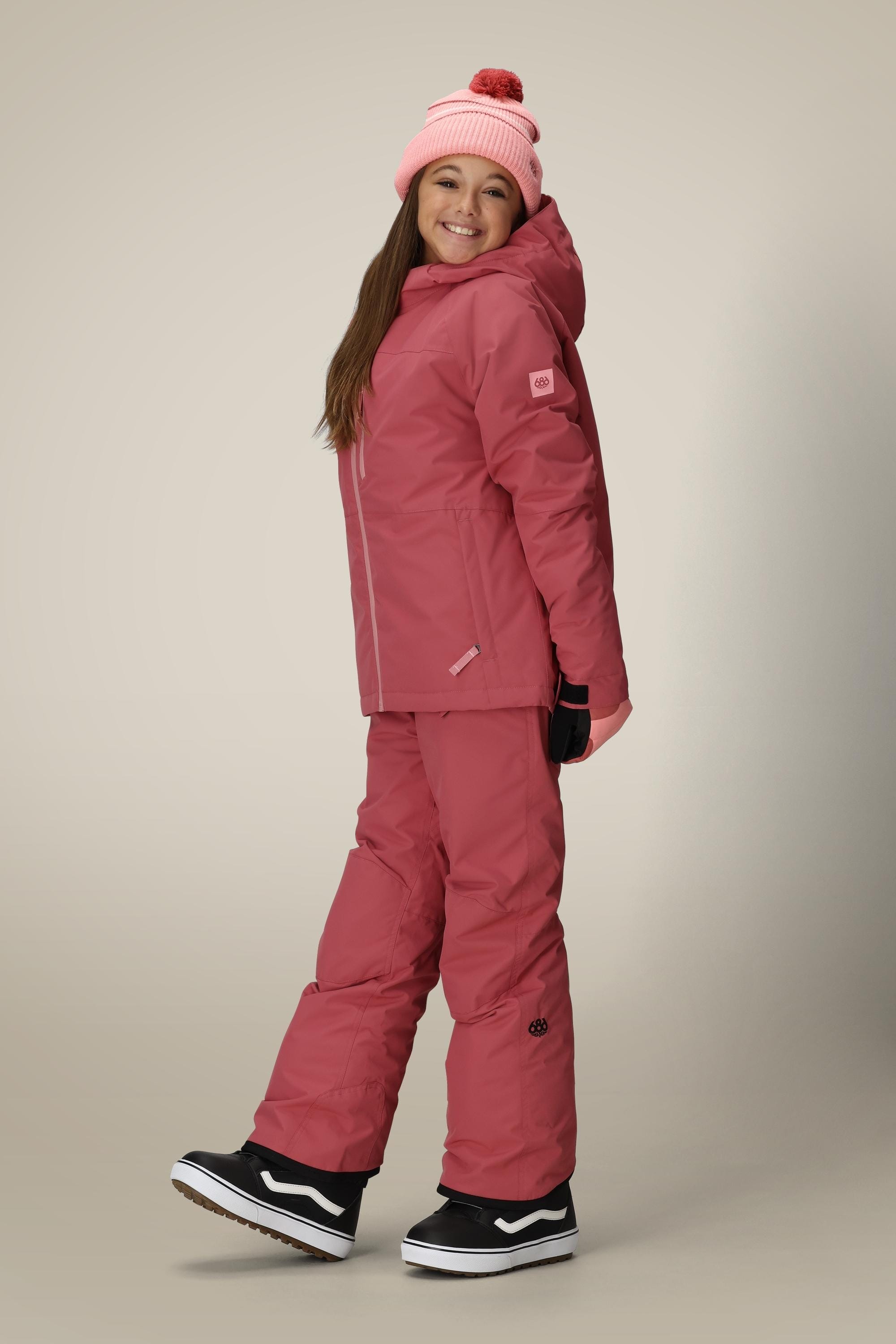 A smiling child models the 686 Girls Hydra Insulated Jacket by 686, paired with pink snow pants, a light pink pom-pom beanie, black gloves, and black boots—posing against a neutral background.