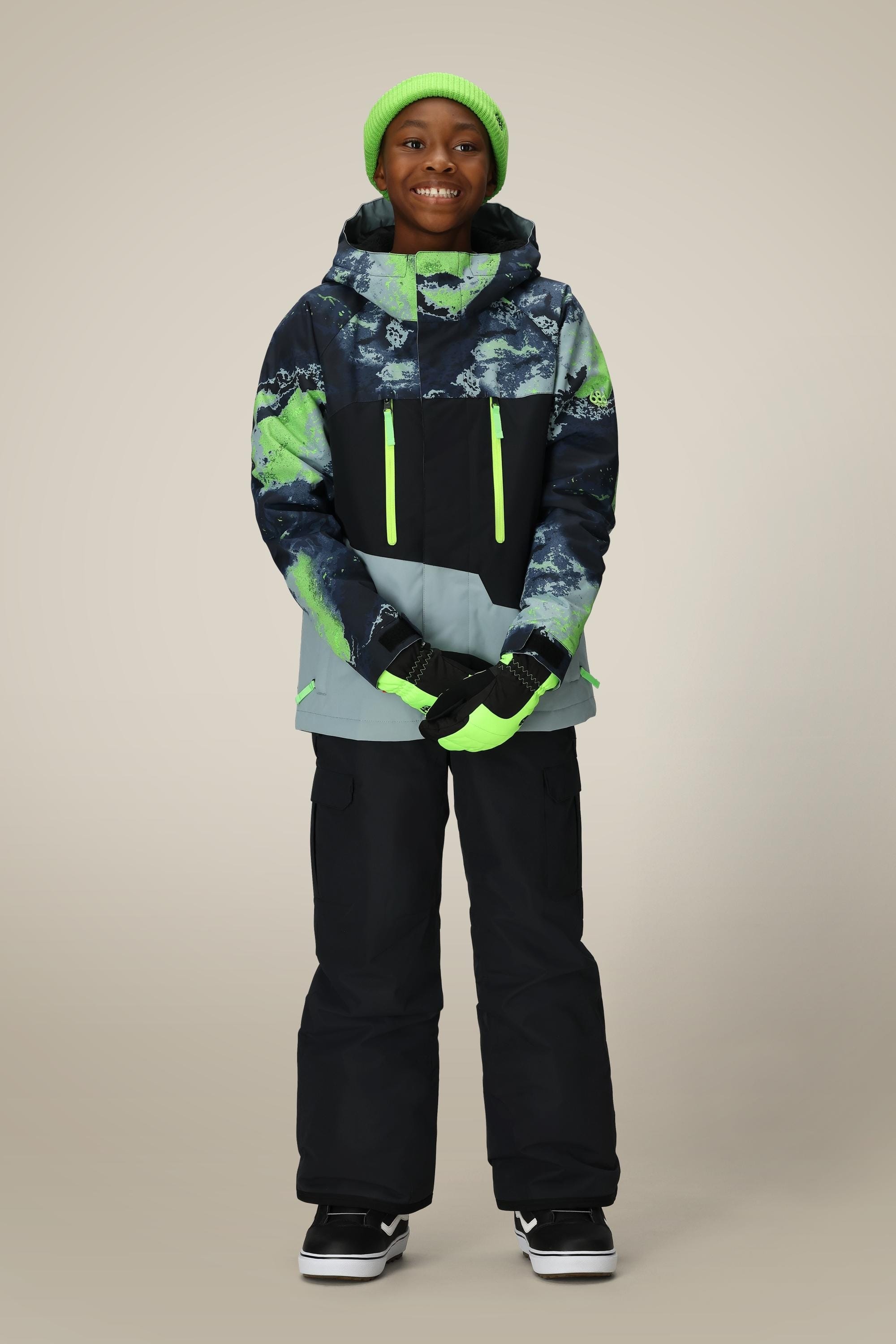 A smiling child models the 686 Boys Infinity Cargo Insulated Pant by 686, paired with a green beanie, colorful patterned winter jacket, gloves, and black sneakers against a plain light background.