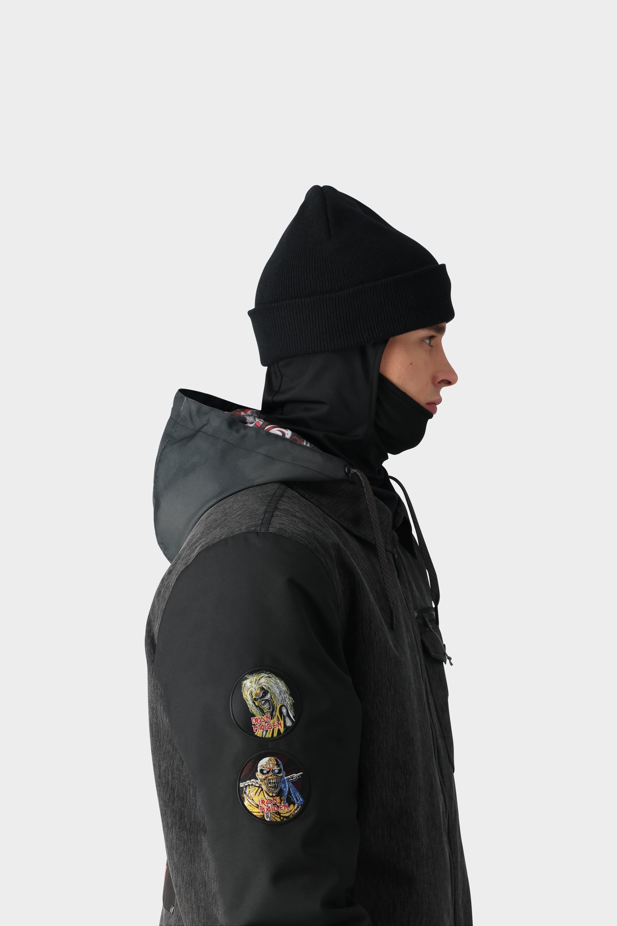 A person in profile wears a black beanie, face covering, and the 686 x Iron Maiden Mens Insulated Battle Jacket by 686 with two colorful embroidered sleeve patches, set against a plain light background.