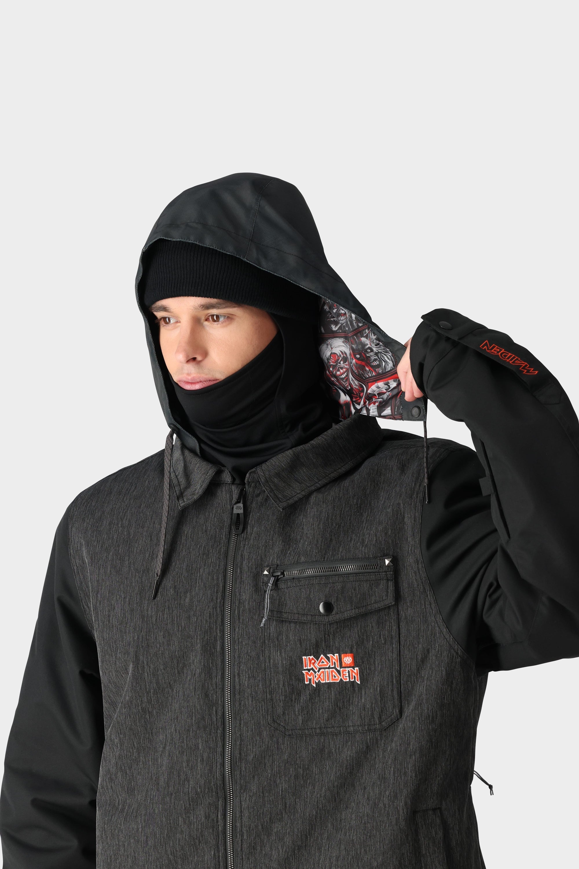A person in the 686 x Iron Maiden Mens Insulated Battle Jacket pulls back their hood to reveal a patterned lining. The black, waterproof, limited edition 686 jacket features red text on the chest and sleeve.