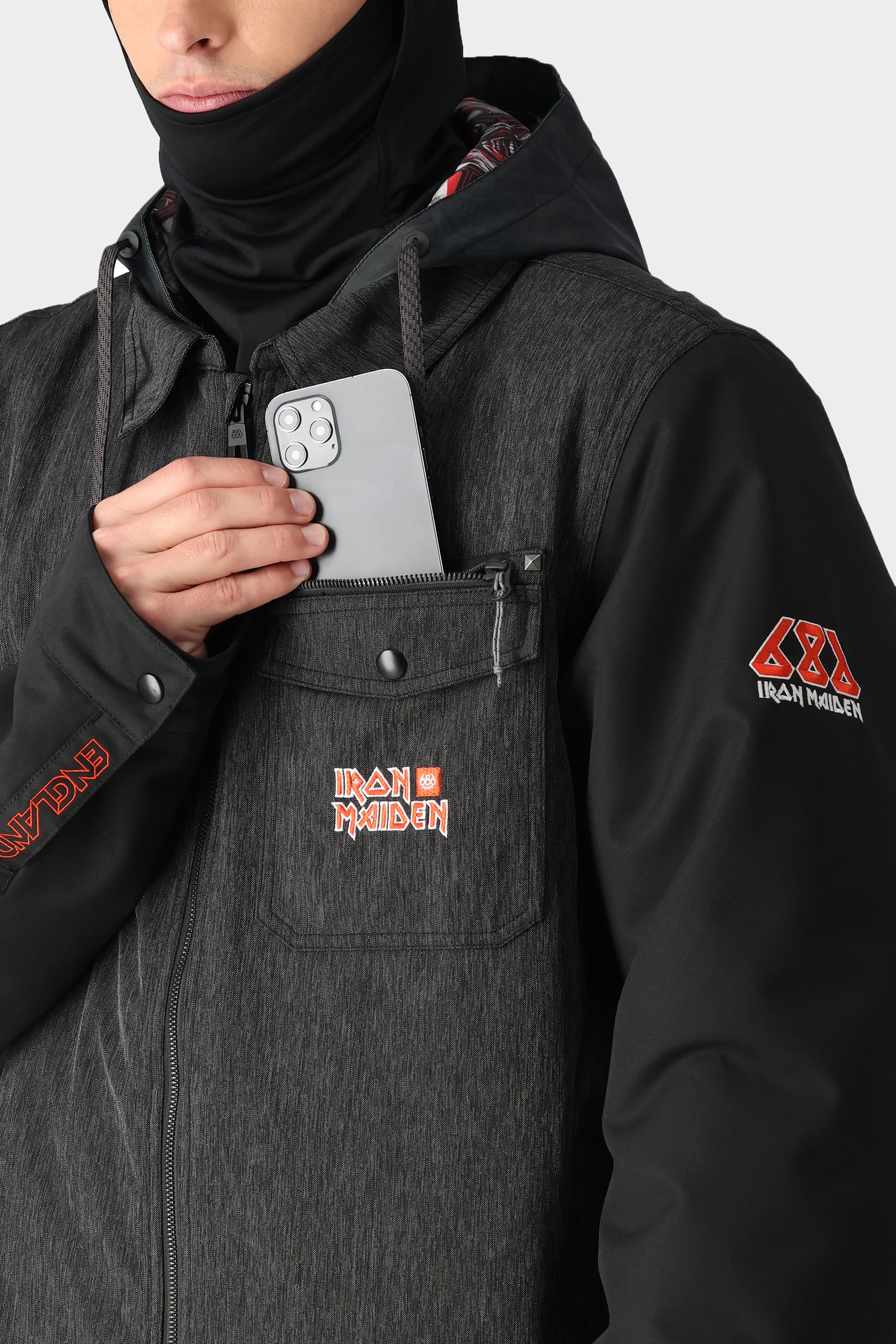Someone wearing the 686 x Iron Maiden Mens Insulated Battle Jacket in dark grey, featuring IRON MAIDEN logos, slips a silver smartphone into its chest pocket. The waterproof jackets hood and a black balaclava partially cover their face.