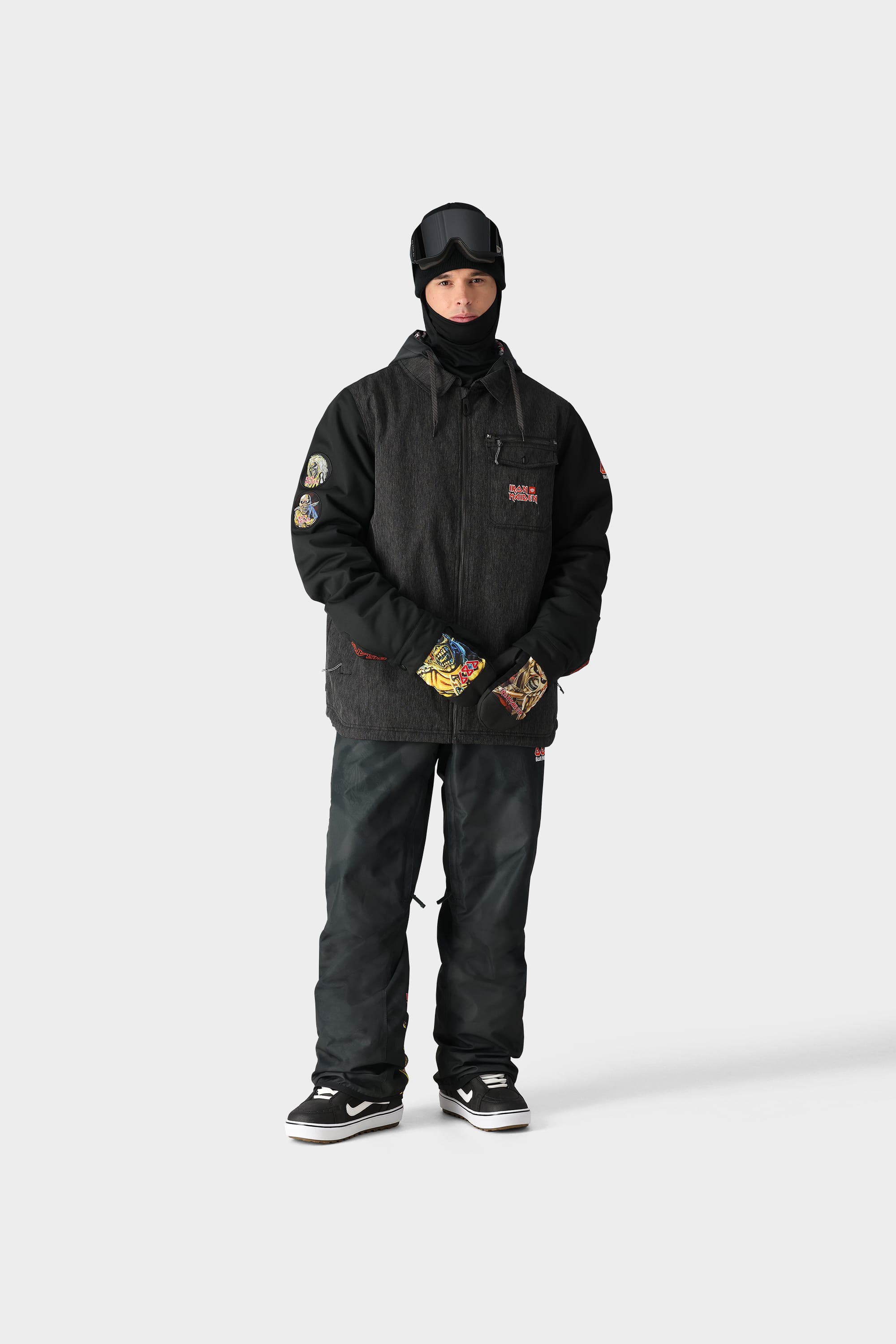 A person in the 686 x Iron Maiden Mens Insulated Battle Jacket, dark pants, gloves, black sneakers, a black helmet, and a face covering stands against a plain light background.