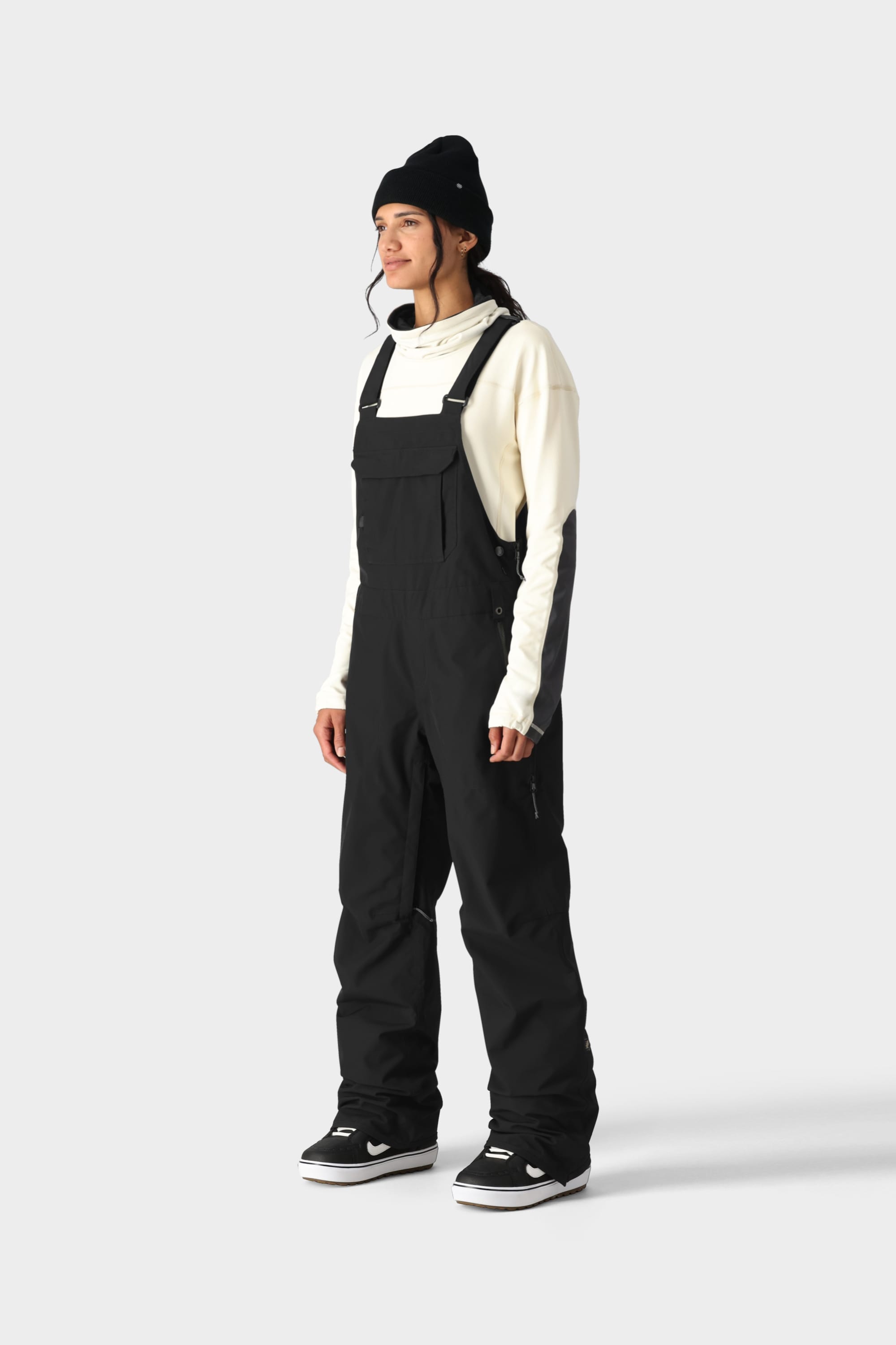 A person faces left against a light grey background, wearing a white hoodie, black beanie, black sneakers with white soles, and the 686 Womens Black Magic 20K Bib by 686 with functional pocketing.