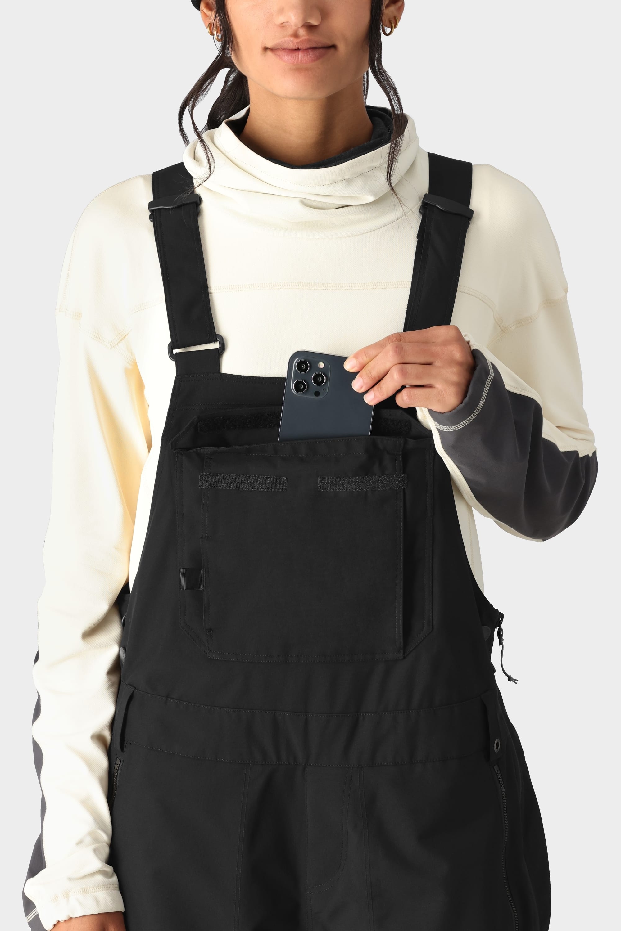 A person wearing a cream and grey long-sleeve top with the 686 Womens Black Magic 20K Bib by 686 is pocketing a smartphone into the bib’s chest pocket, set against a plain, light-colored background.