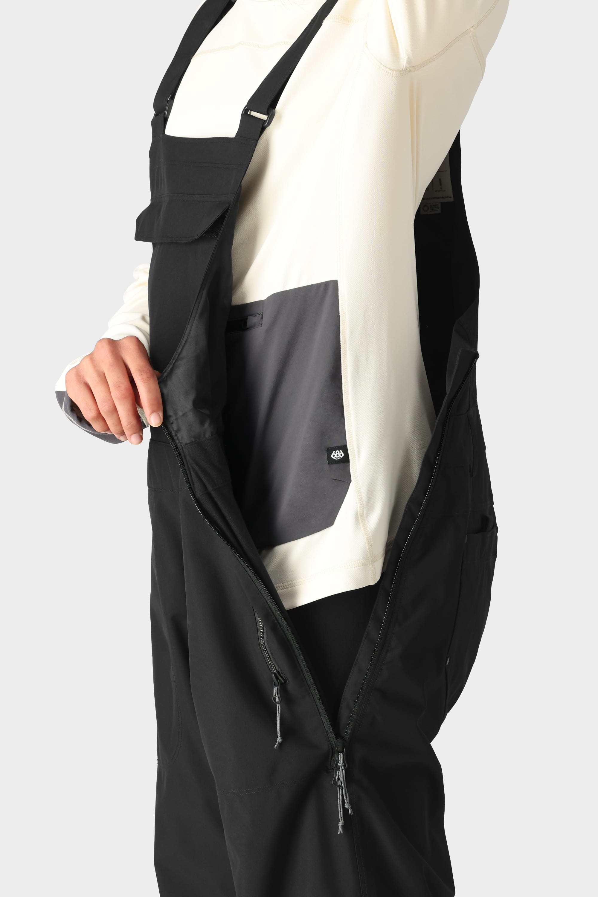 A person wears the 686 Womens Black Magic 20K Bib with a side zipper open, showing grey inner pocketing, layered over a long-sleeve cream shirt. The look is set against a plain light background. Brand: 686.