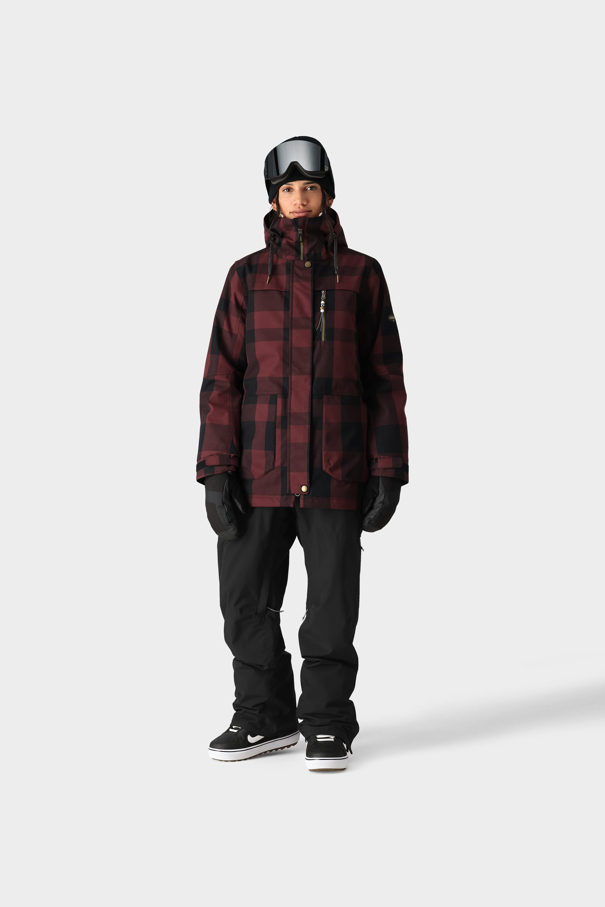 A person wears the 686 Womens Black Magic 20K Bib from 686, a red and black plaid insulated winter jacket, black gloves, snow boots, and a black helmet with ski goggles, standing against a plain light grey background.