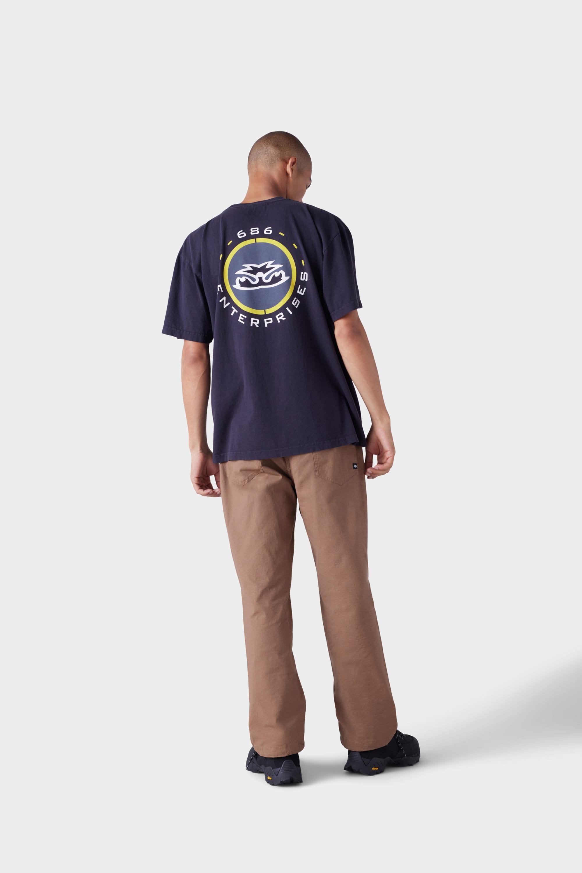 A person stands facing away, wearing the 686 Mens Jib 2001 Badge Short Sleeve Tee in navy with a circular yellow and white back graphic, brown pants, and black shoes, against a plain light grey background.