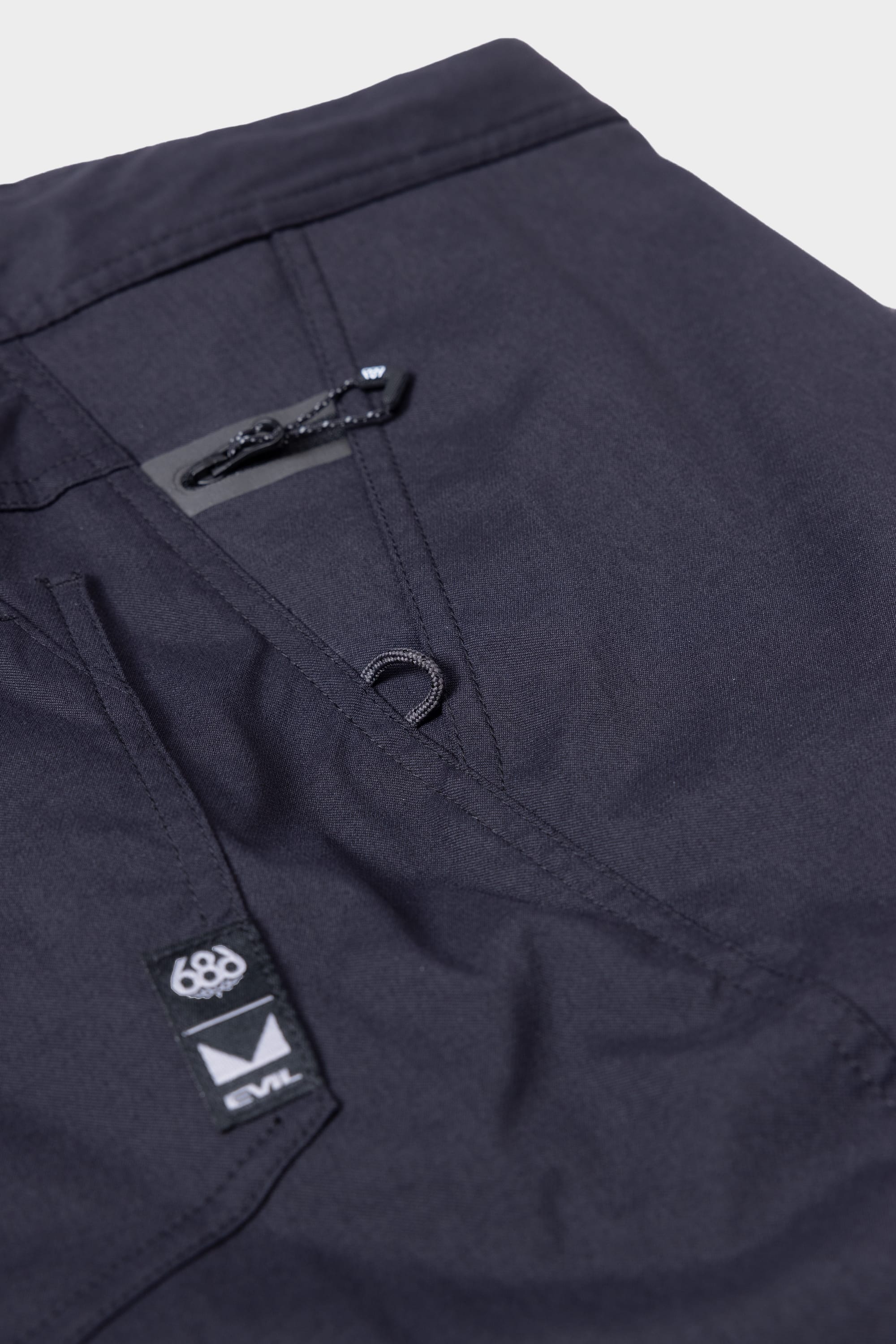 Close-up of the 686 Mens Platform Bike Pant - Relaxed Fit showing a zipper pocket, loop detail, and two small fabric labels featuring the 686 and EVIL logos on its durable, slightly textured fabric.