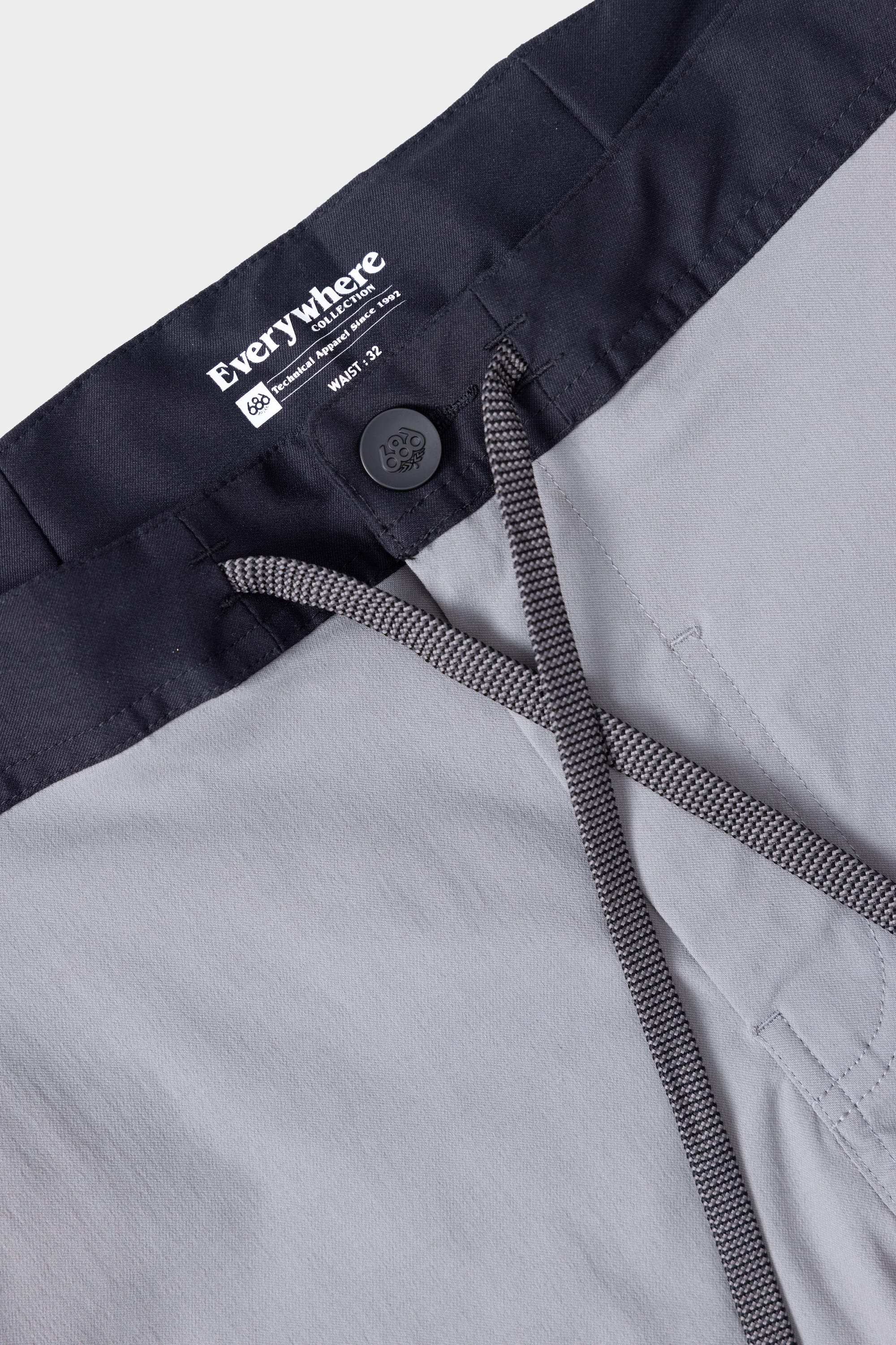 Close-up of 686 Mens Platform Bike Pant - Slim Fit in light grey, featuring a black waistband, black button, and grey drawstring. The inside label shows the 686 brand and details on size and manufacturing.