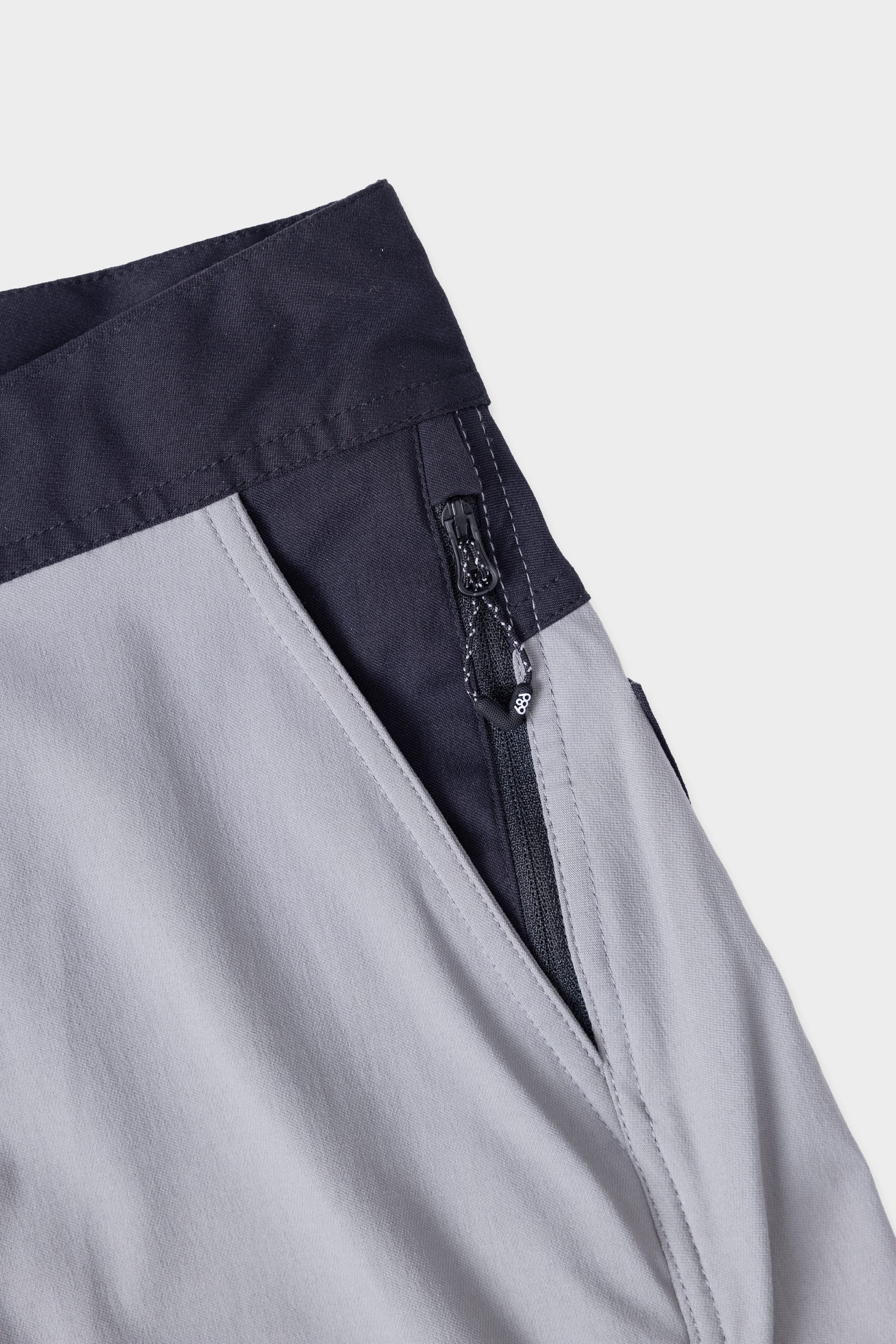 Close-up of the 686 Mens Platform Bike Pant - Slim Fit, showing light grey fabric, dark grey waistband, a visible zipper pocket with pull, and prominent stitching details.