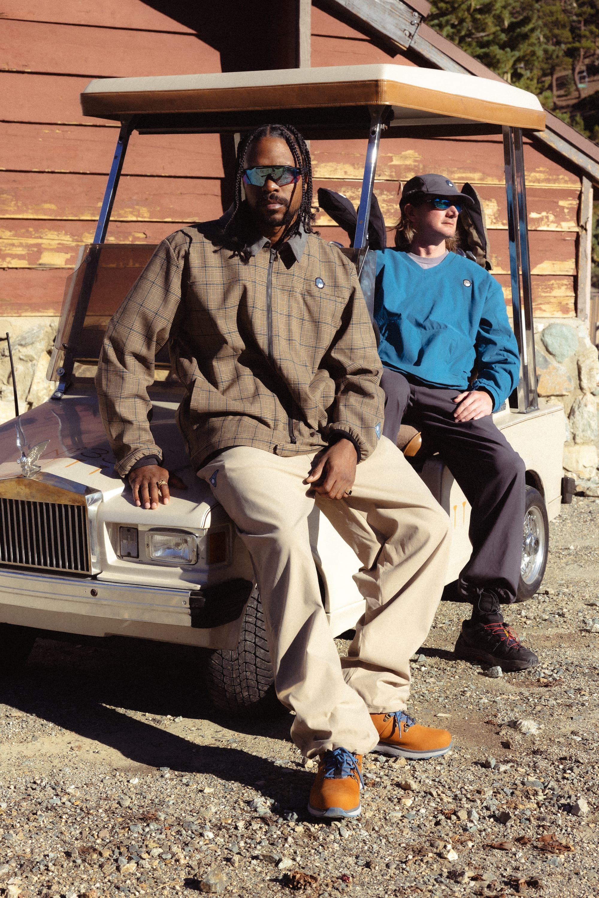 Two people sit on a golf cart outside a rustic wooden building. In front, a person wears sunglasses, a brown plaid shirt, and khaki pants; behind them, someone sports the 686 x Malbon Mens Caddie Performance Jacket by 686 with black pants.