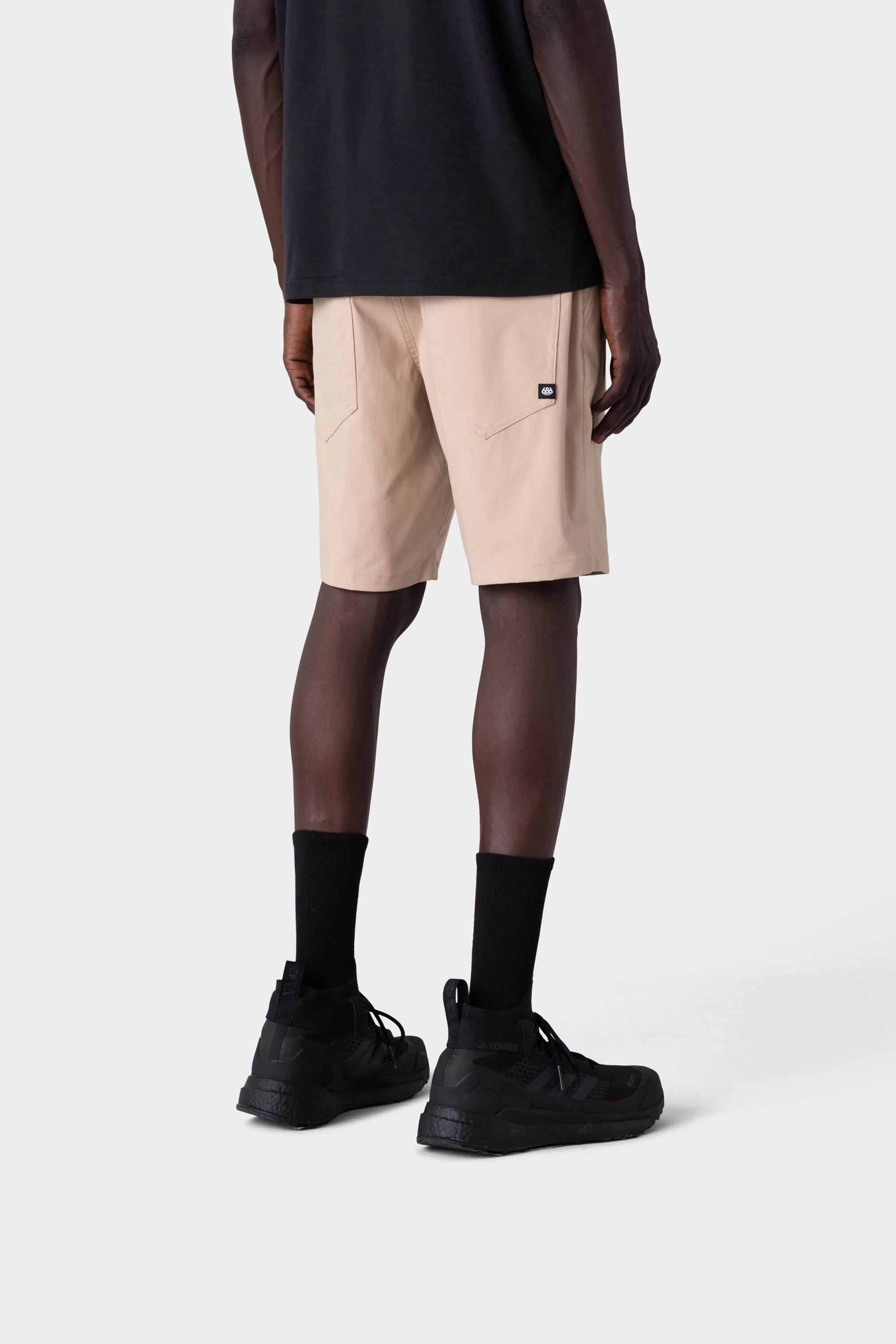 A person stands facing away, wearing a black T-shirt and 686 Mens Everywhere Hybrid Short by 686 with back pockets, paired with black crew socks and athletic shoes against a plain light gray background.