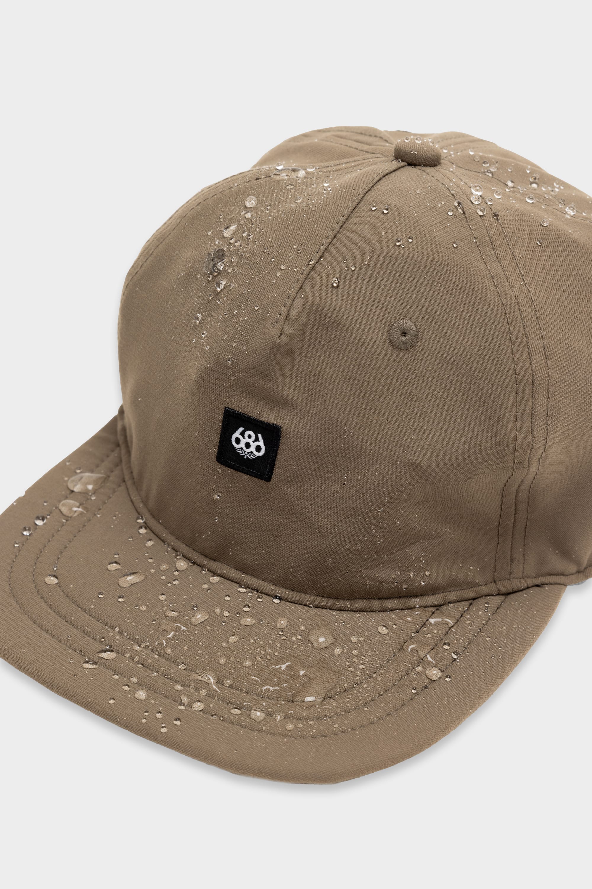 The 686 Packable Everywhere Hat by 686 is a khaki baseball cap shown with water droplets on its surface and a black rectangular front patch featuring a white circular symbol, set against a plain light background.