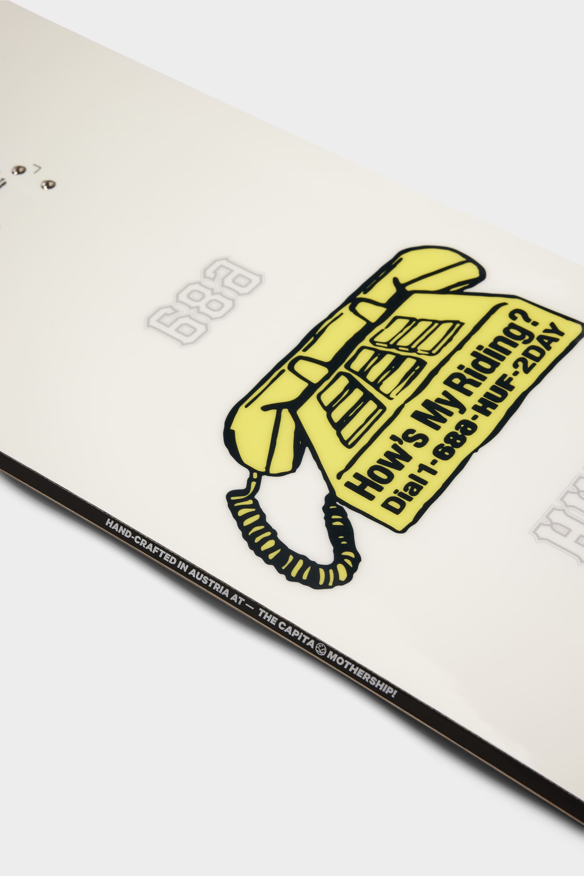 Close-up of the 686 HUF Outerspace Living Snowboard by 686, featuring a glossy white finish, yellow cartoon telephone graphic with How’s My Riding? Dial 688-HUT-2DAY, embossed numbers, edge text, and a playful freestyle shape for tricks.