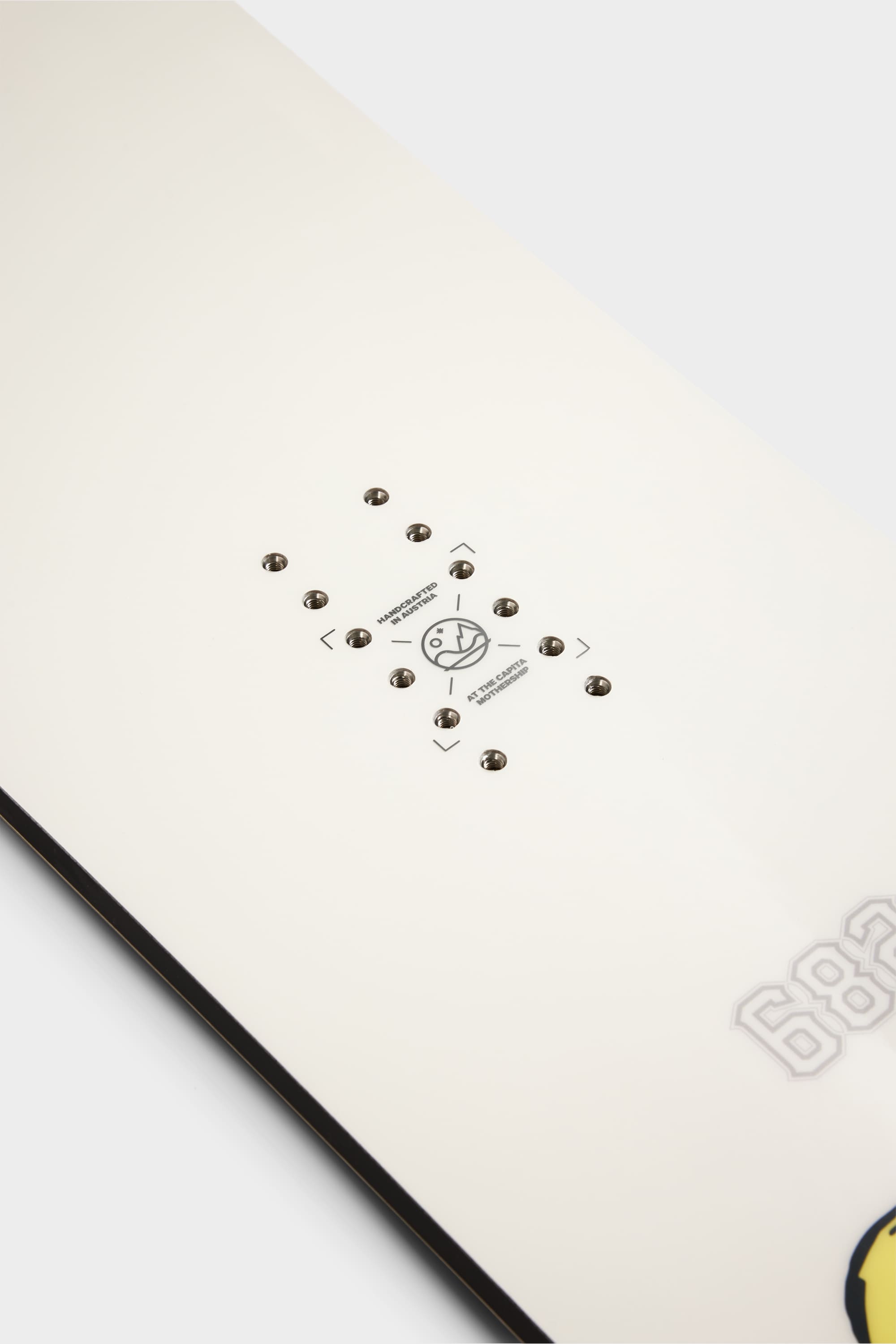 Close-up of the 686 HUF Outerspace Living Snowboard’s mounting area, showing multiple screw holes and alignment markings on a white surface, highlighting its all-terrain camber versatility.
