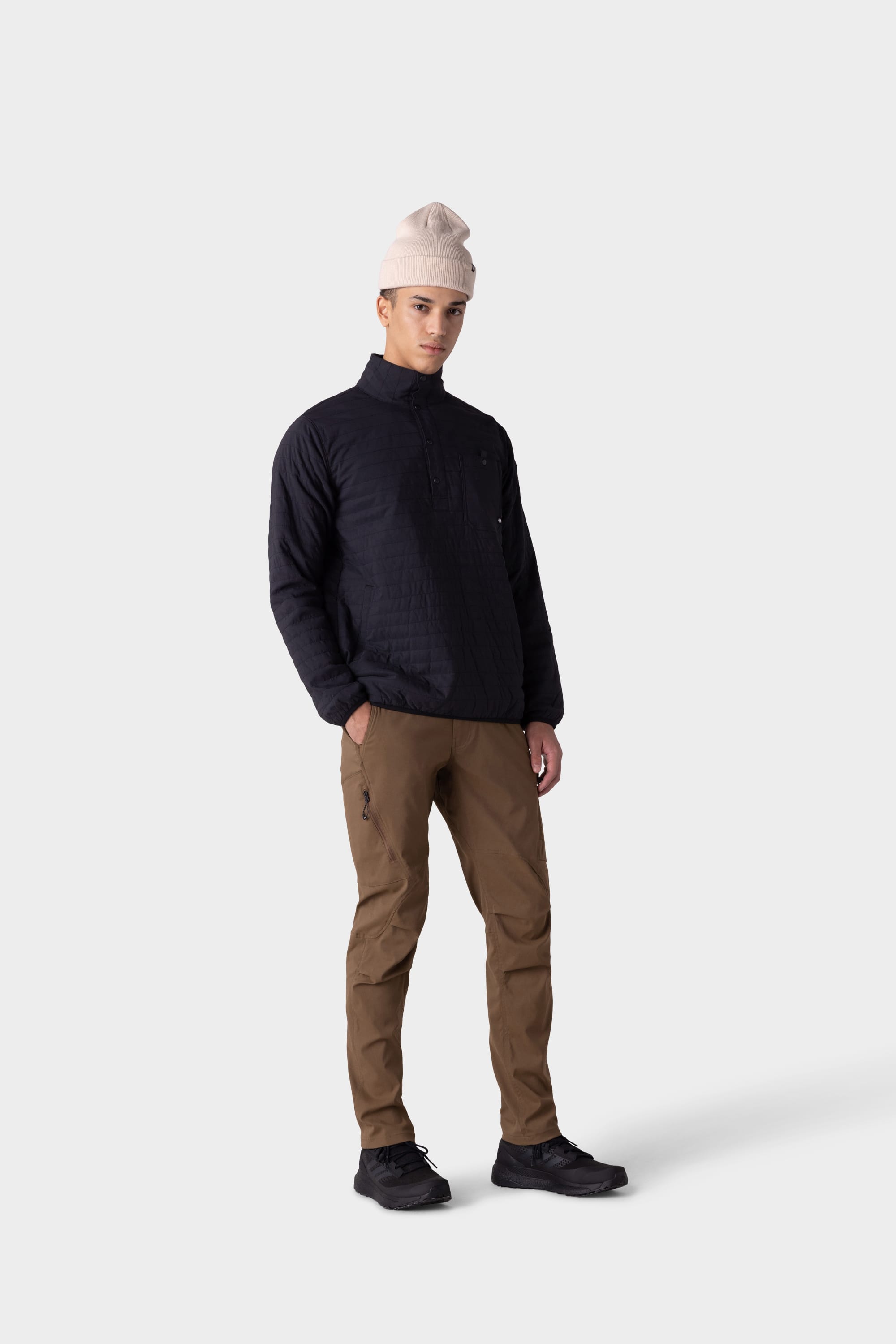 A person stands facing forward, wearing a light beige beanie, a black quilted jacket, 686 Mens Anything Cargo Pant - Slim Fit by 686, and black shoes against a plain light grey background.