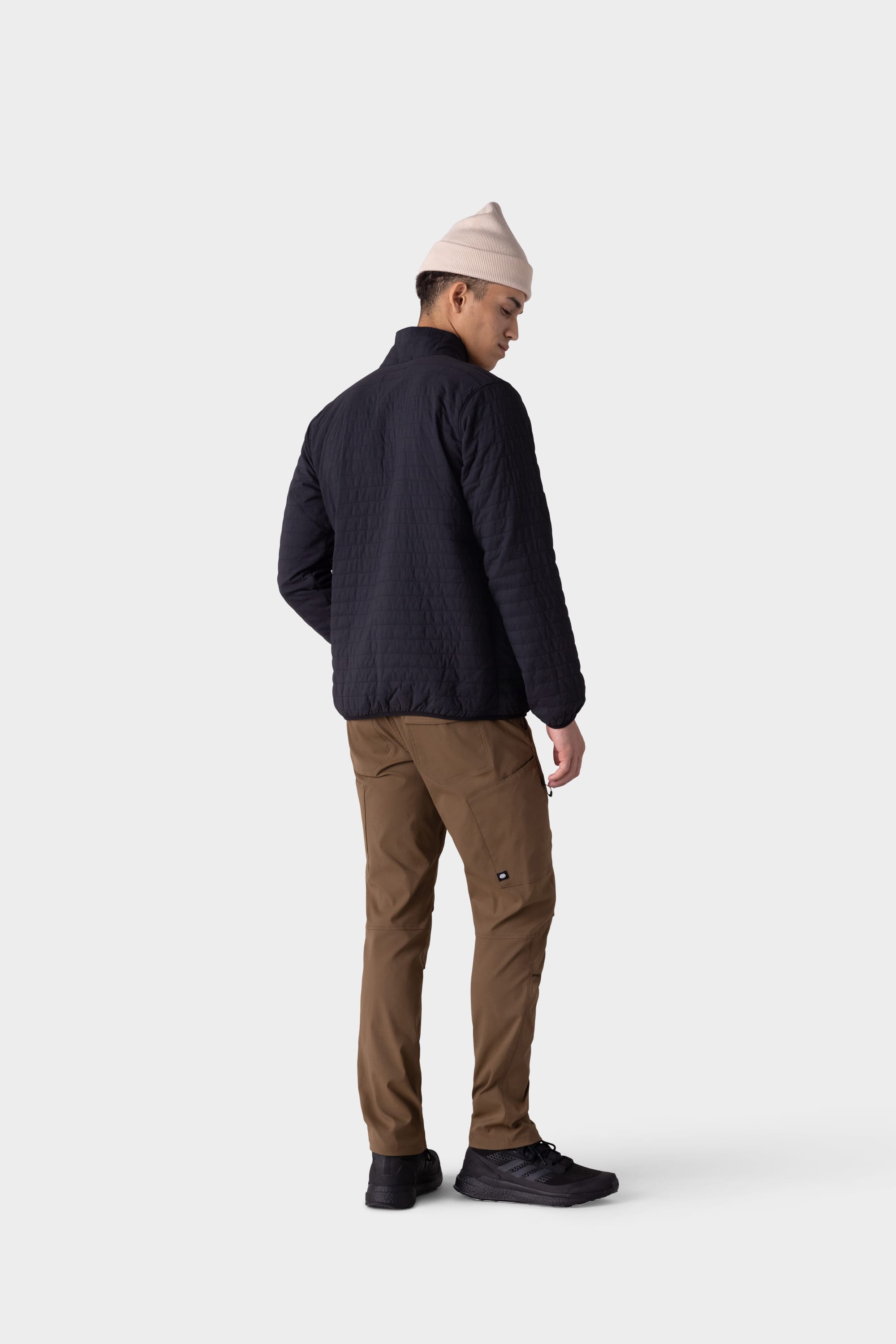 A person wearing a light beige beanie, black quilted jacket, 686 Mens Anything Cargo Pant - Slim Fit by 686, and black sneakers stands facing away, looking over their left shoulder against a plain light background.