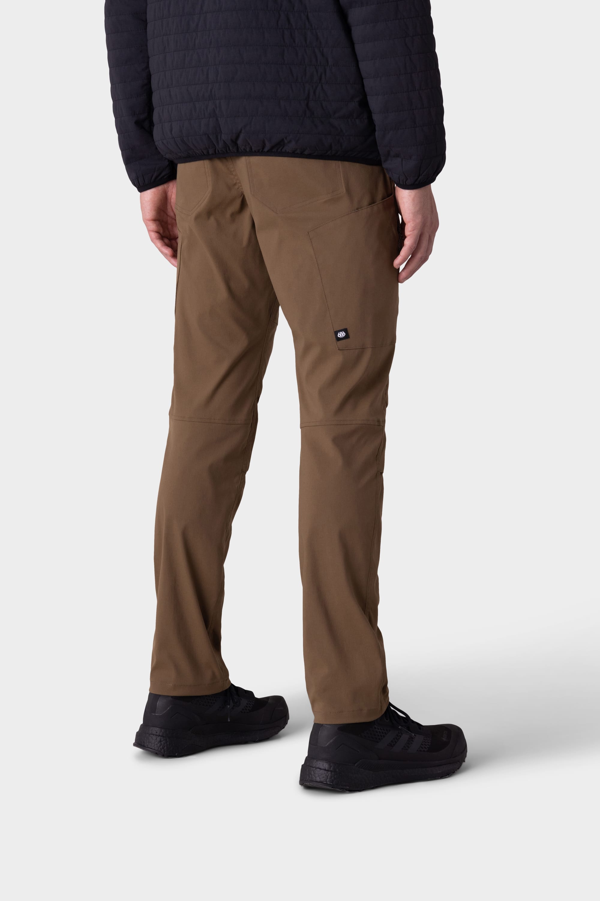 A person faces away, wearing 686 Mens Anything Cargo Pant - Slim Fit by 686 in brown with a 13-pocket design, paired with a black quilted jacket and black sneakers, against a plain light background.