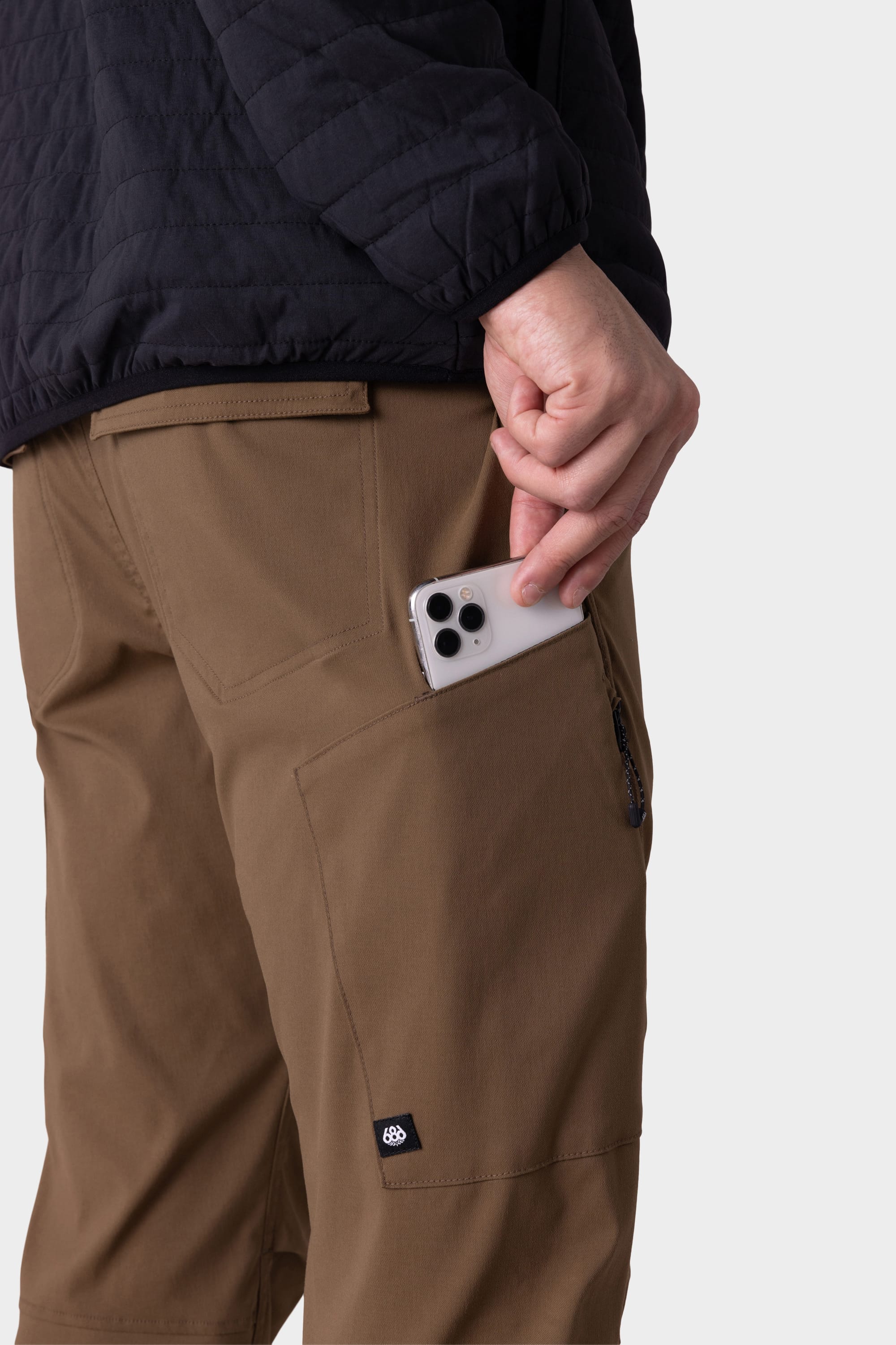 A person in 686 Mens Anything Cargo Pant - Slim Fit by 686, wearing a black jacket, slips a triple-lens smartphone into the zippered side pocket of their breathable, brown 13-pocket technical cargo pants.