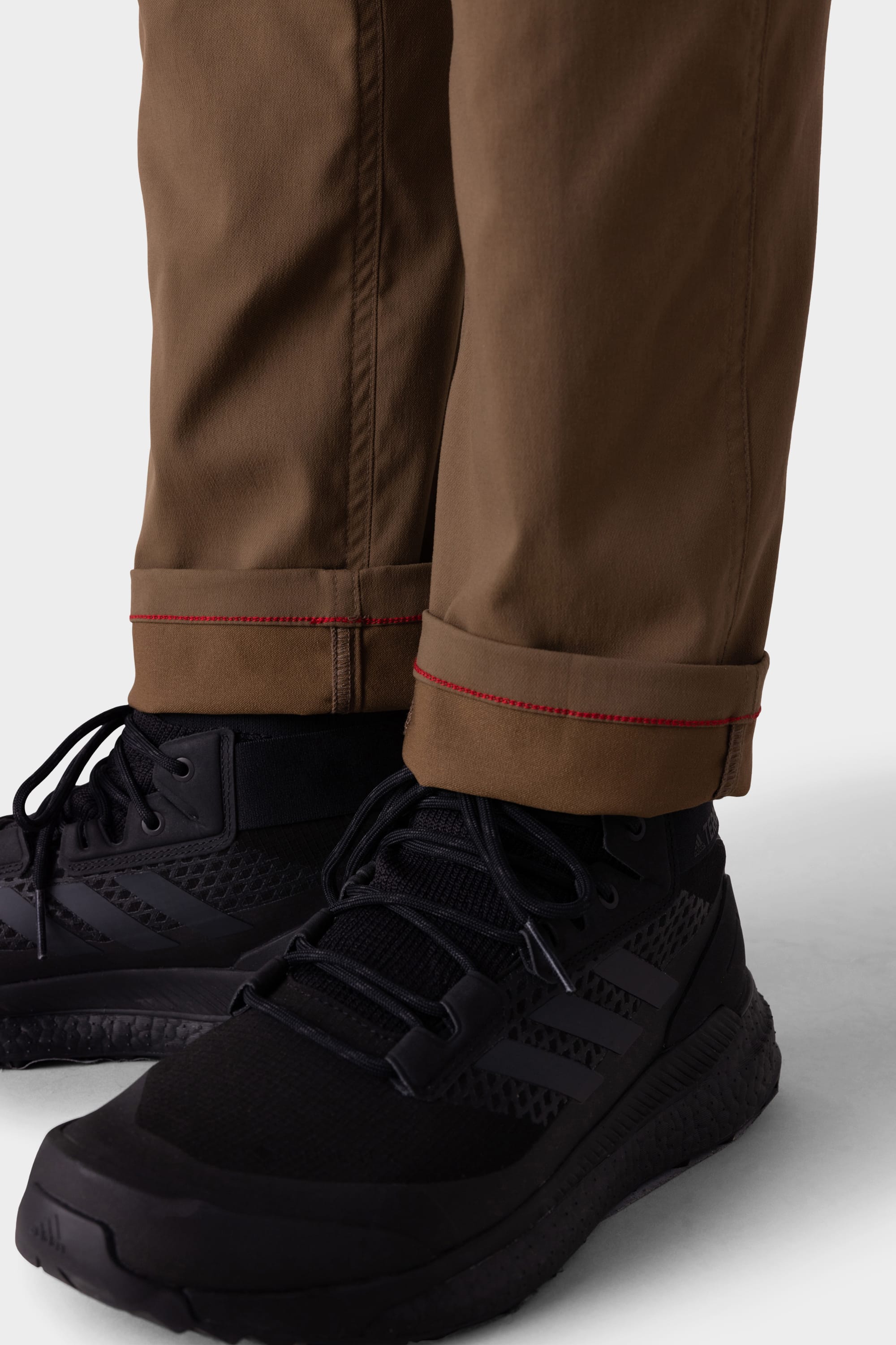 A close-up of a person wearing 686 Mens Anything Cargo Pant - Slim Fit in breathable brown fabric with red-stitched cuffs, paired with black lace-up athletic shoes, set against a light background.