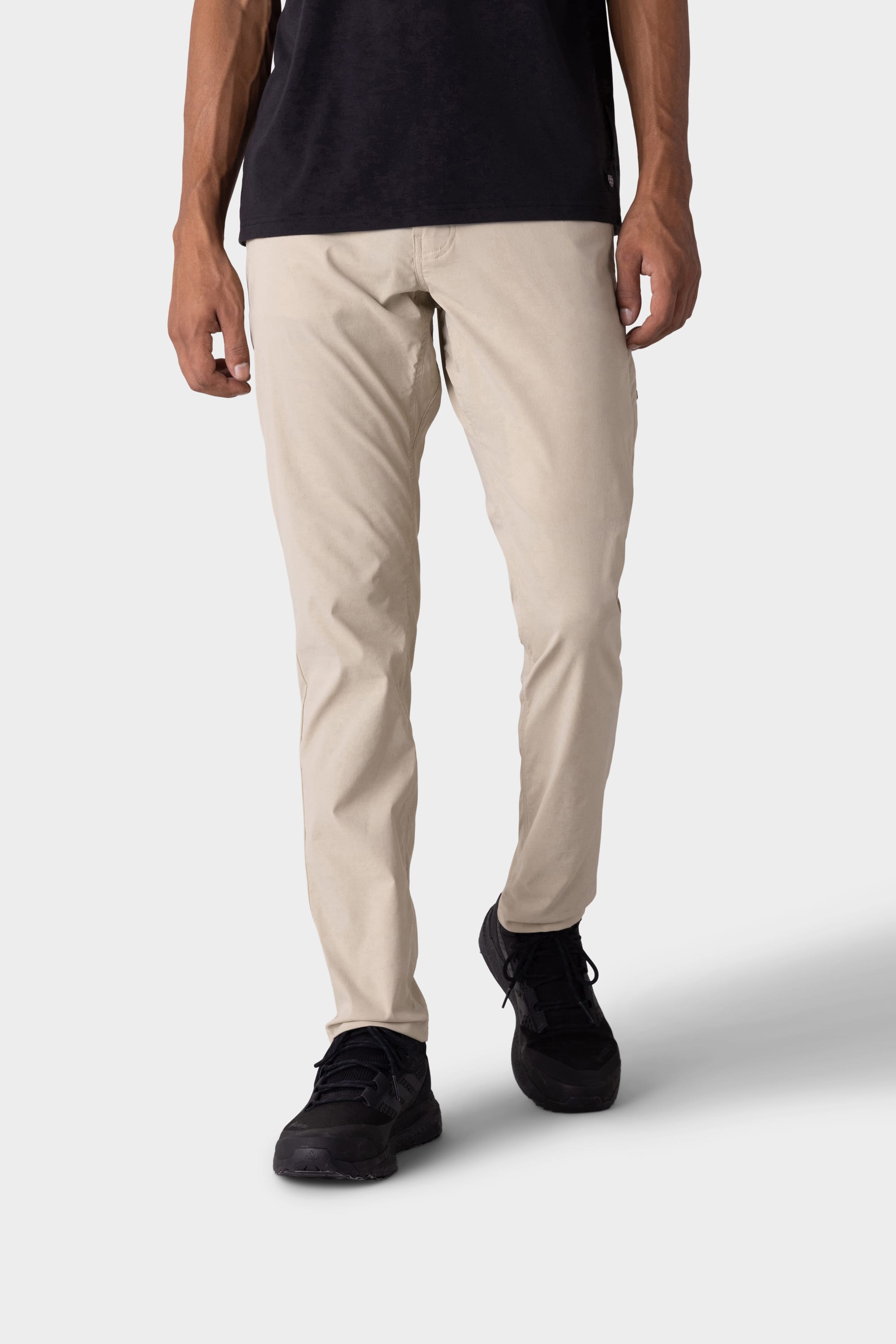 A person stands against a light grey background wearing 686 Mens Everywhere Featherlight Chino Pant - Slim Fit in beige, paired with a black shirt and shoes—ideal lightweight style by 686 for summer comfort and warm weather adventures.