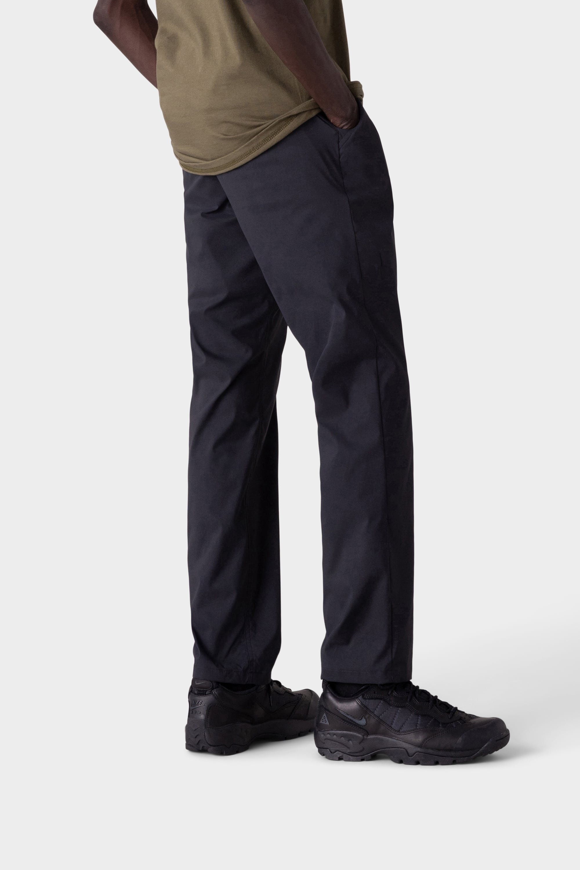 A person stands with hands in pockets, wearing 686 Mens Everywhere Featherlight Chino Pant - Relaxed Fit in black, a khaki t-shirt, and black sneakers against a plain light background. Only the lower body is shown.