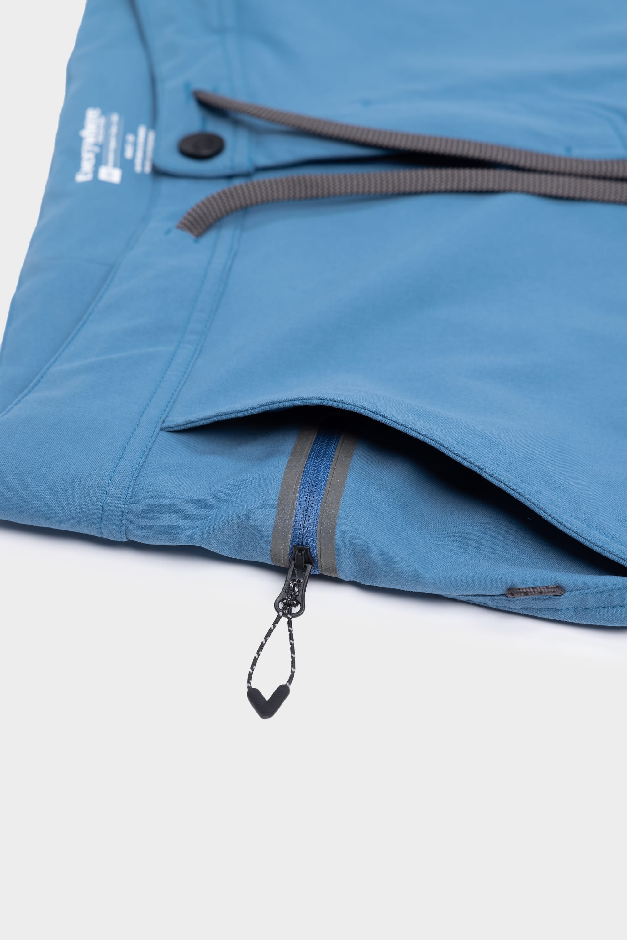 Close-up of the 686 Mens Platform Bike Pant - Slim Fit in blue, showcasing a zippered side pocket with black zipper pull, button closure, and a partially visible waistband against a white background.