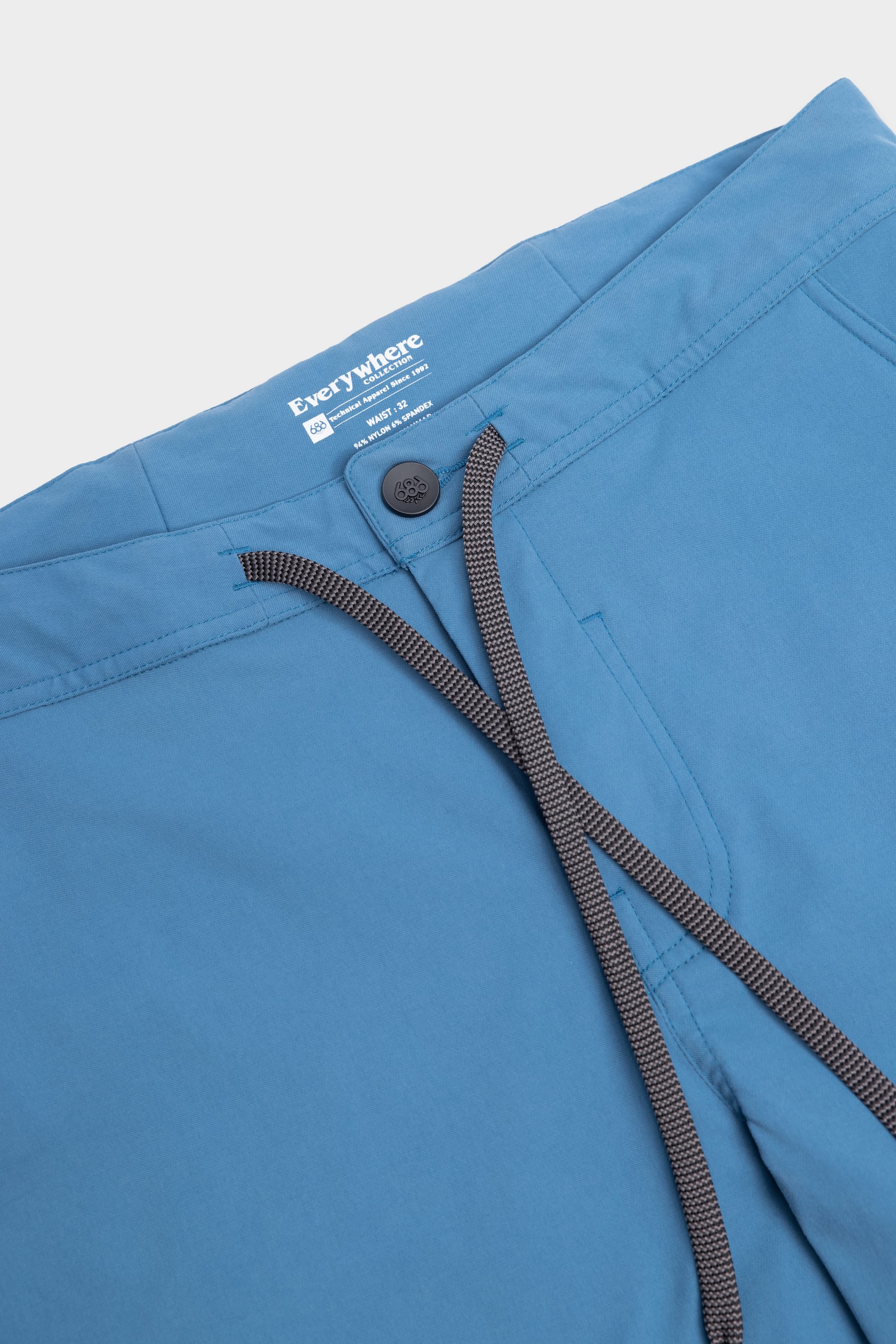 Close-up of 686 Mens Platform Bike Pant - Slim Fit in blue, featuring a black button, zipper fly, and dark grey drawstring. The inner waistband label shows the 686 brand with care and size details.