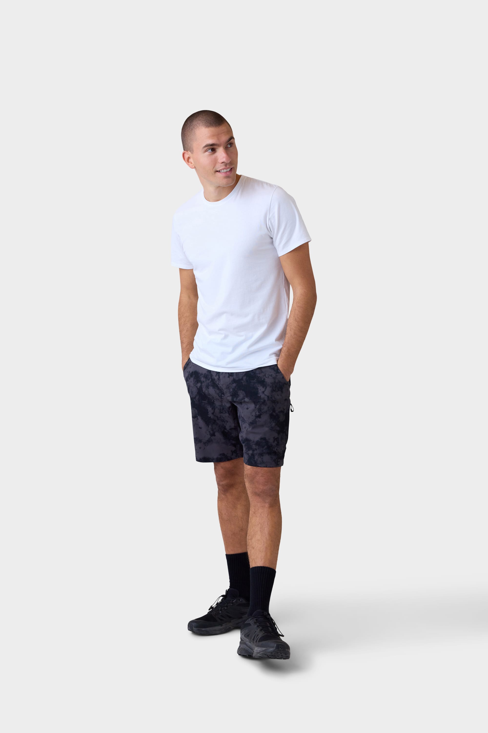 A man wearing a plain white t-shirt, 686 Mens Everywhere Hybrid Short by 686, black socks, and black sneakers stands with his hands in his pockets, looking to the side against a plain light background.