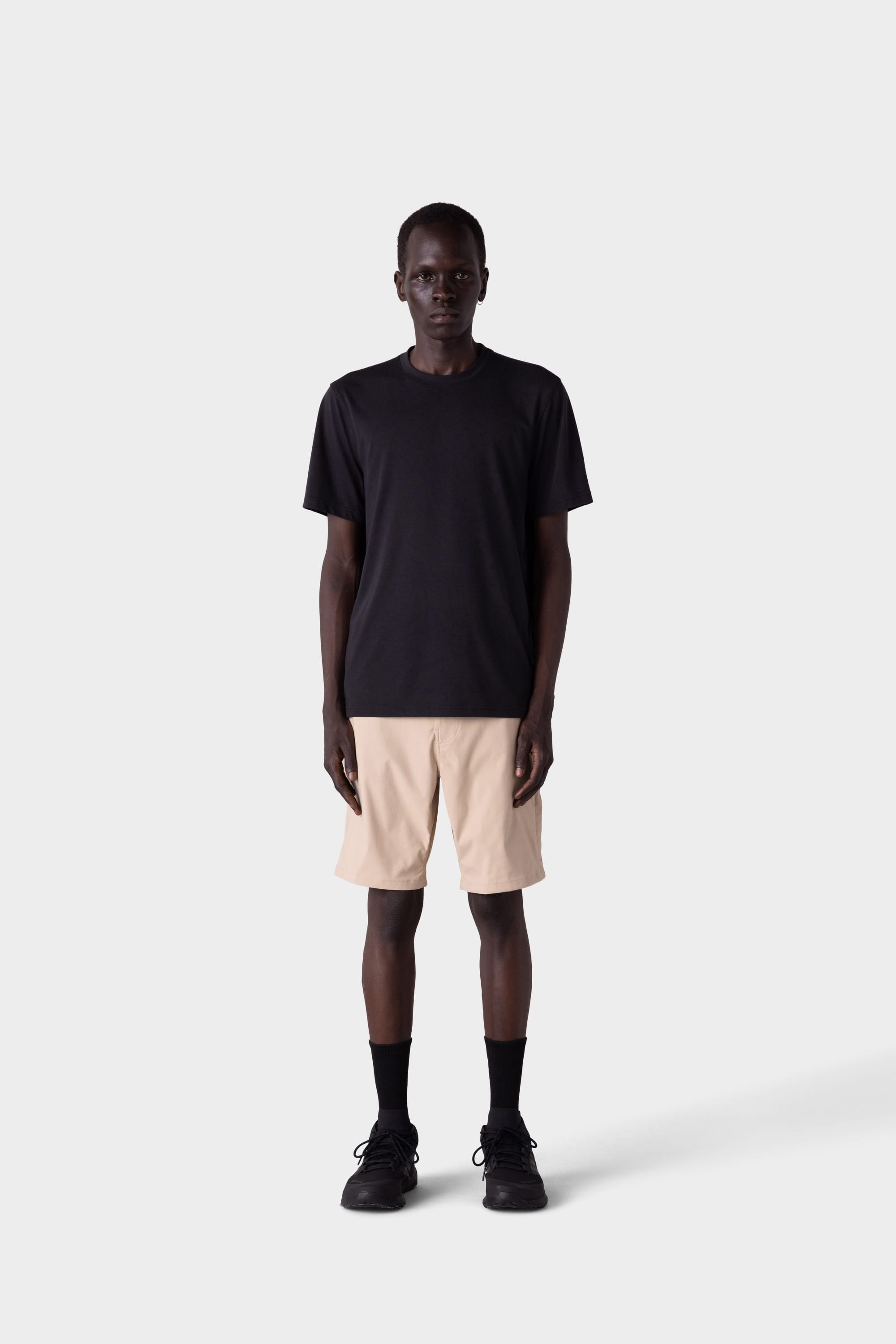 A person stands facing forward against a light gray background, wearing a plain black T-shirt, 686 Mens Everywhere Hybrid Shorts in beige, black socks, and black shoes.