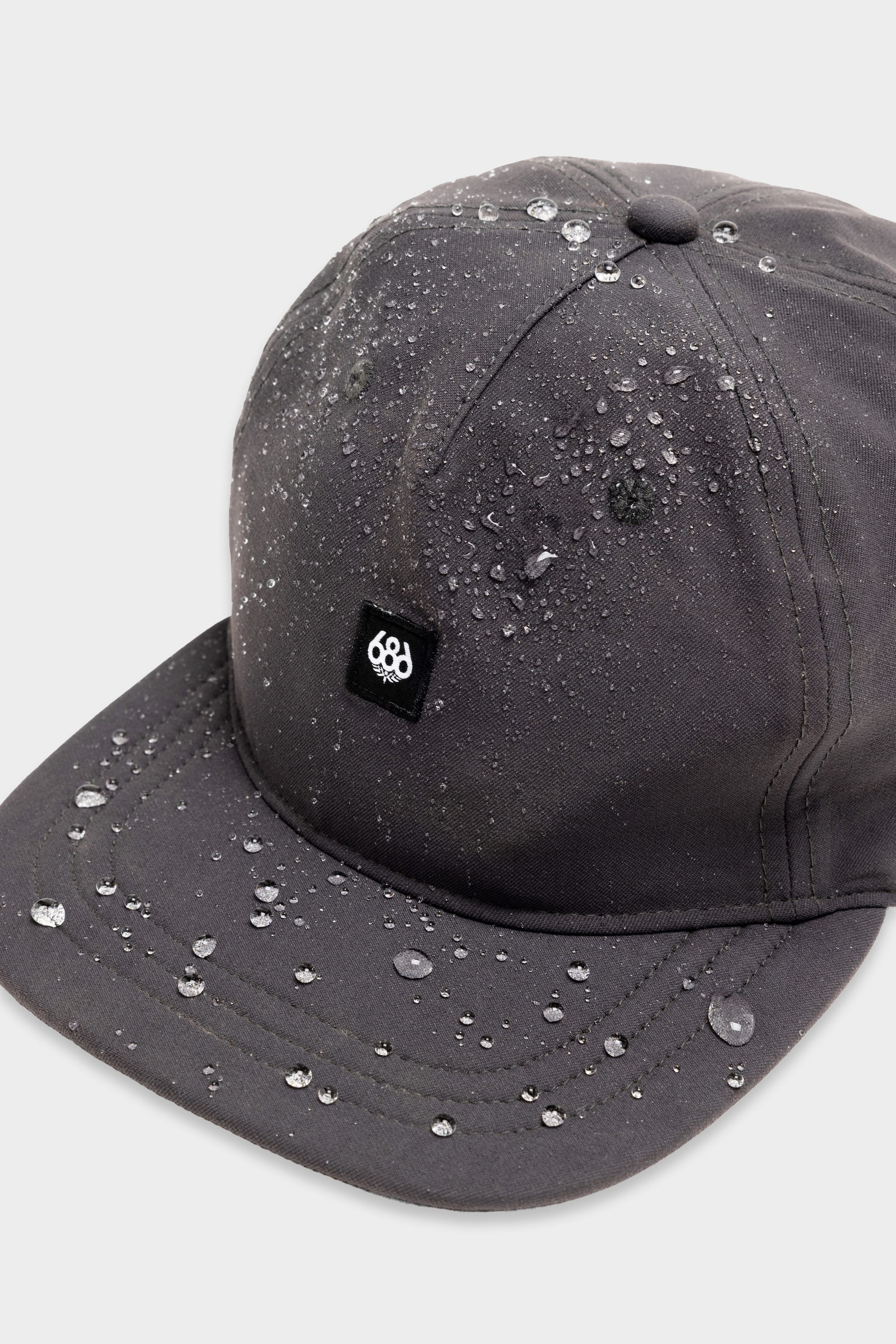 The 686 Packable Everywhere Hat by 686 is a dark gray baseball cap with water droplets on its surface and a small black and white patch logo on the front, shown against a plain light background.
