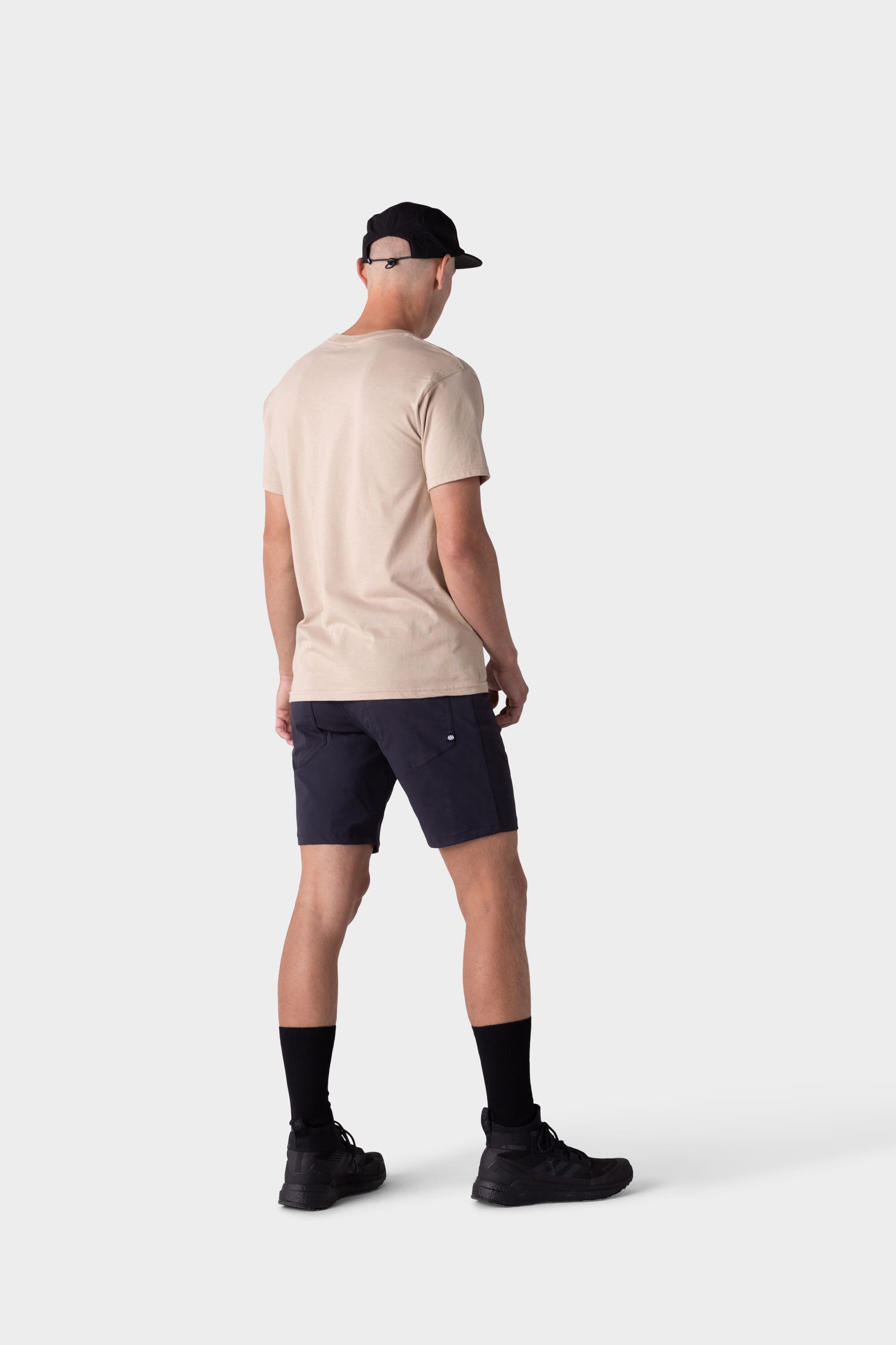 A person wearing a beige t-shirt, 686 Mens Everywhere Hybrid Short by 686, black socks, sneakers, and cap stands facing away on a plain light background.
