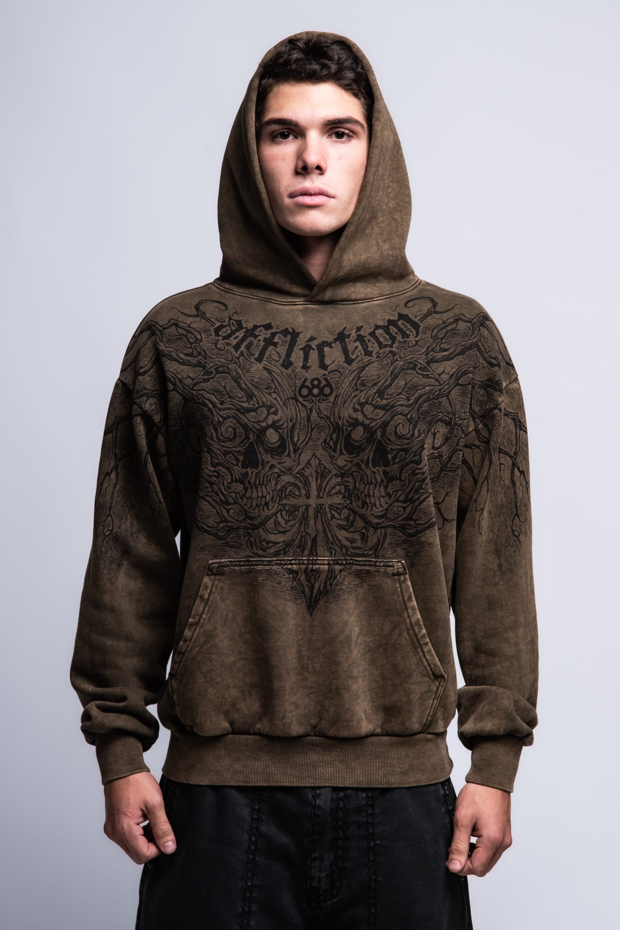 A young man faces forward against a plain light-gray background, wearing the 686 Affliction Natural Rot Pullover Hoody by 686, a brown men’s sweatshirt featuring black intricate designs and "Affliction" on the chest.